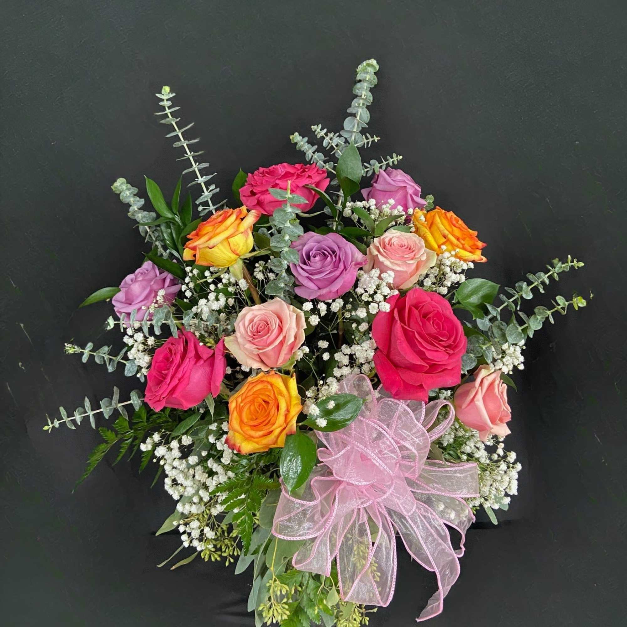 Mixed arrangement of pink, peach, lavender, and yellow roses with white filler flowers and a pink ribbon bow