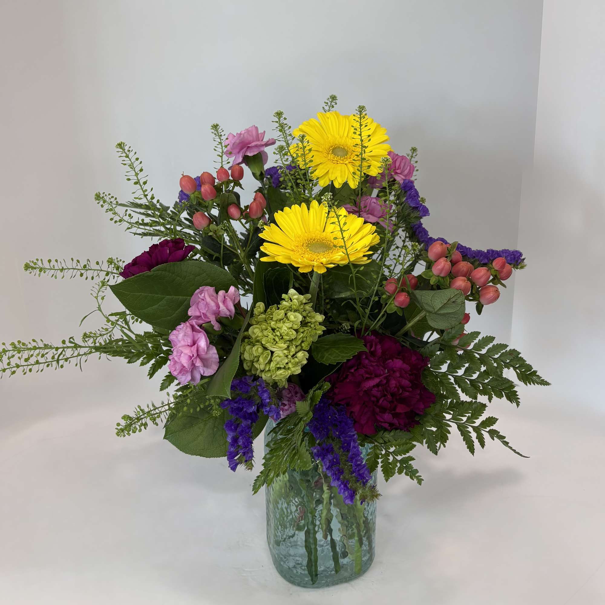 Mixed bouquet of yellow gerbera daisies, pink blooms, and purple flowers in a glass vase