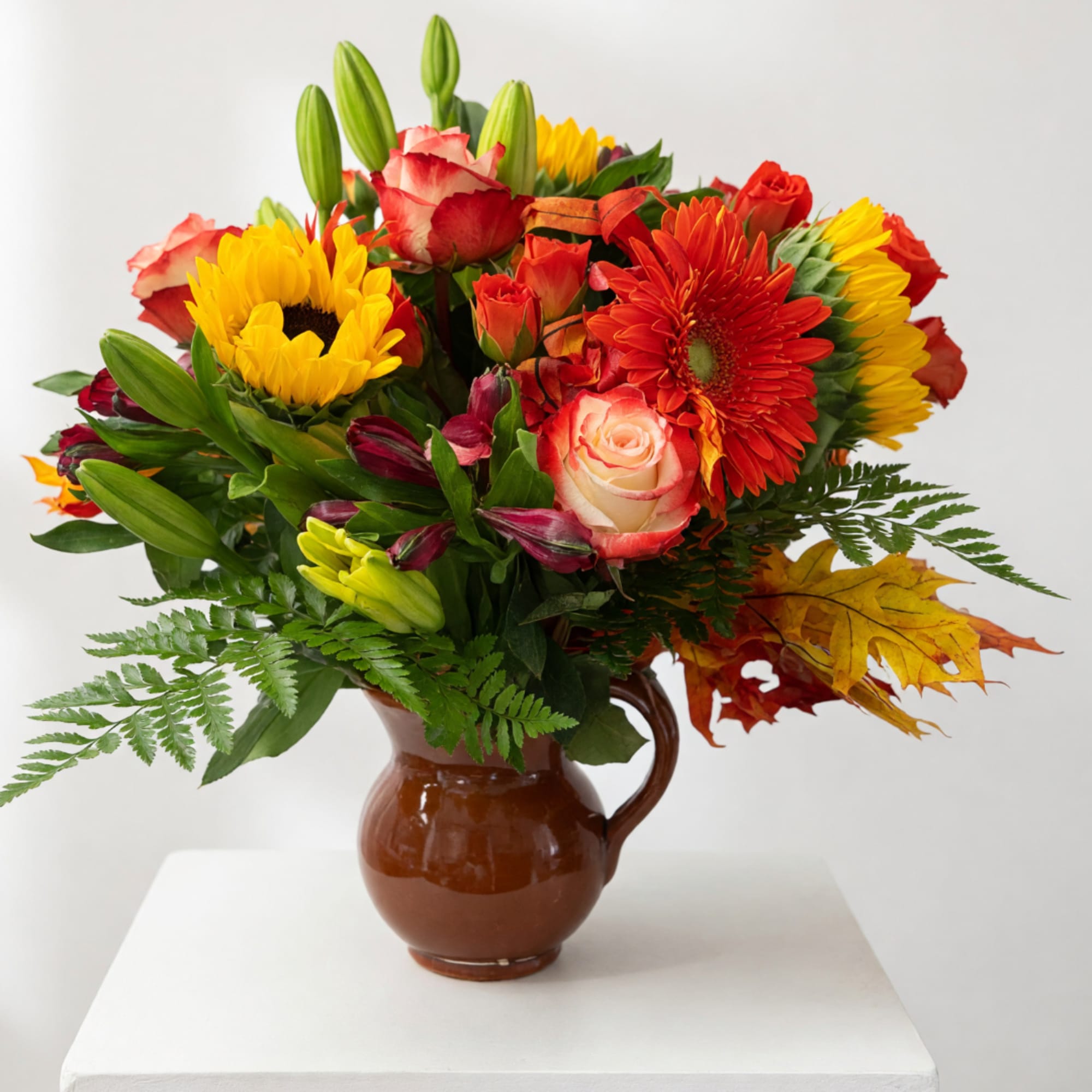 Mixed bouquet of sunflowers, roses, lilies, and gerbera daisies in a brown pitcher vase