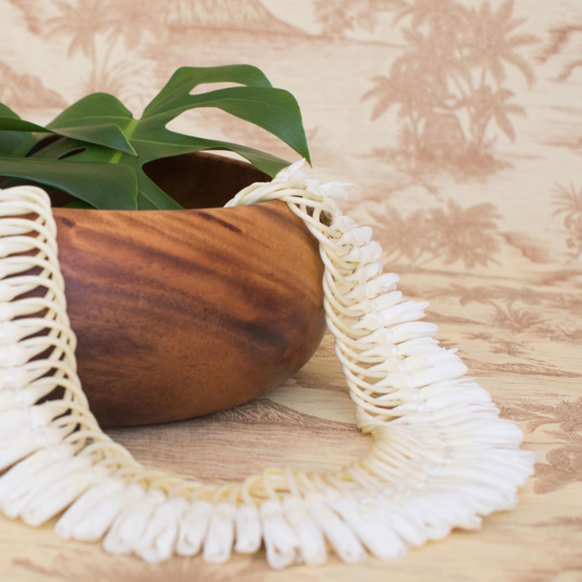 White shell lei draped over a wooden bowl with large green leaves