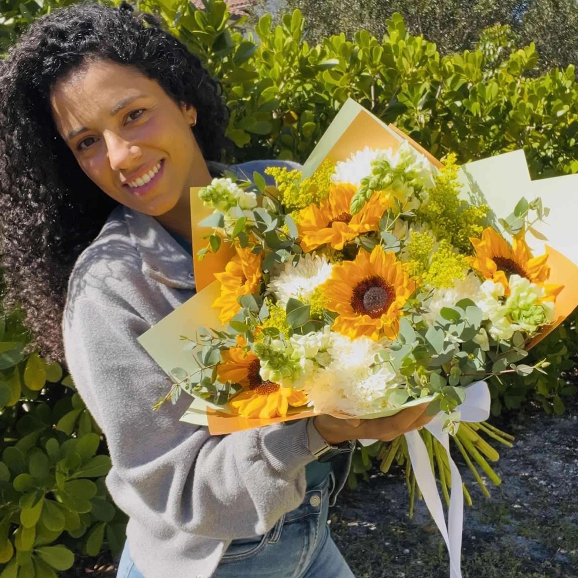 Person holding a large bouquet of yellow sunflowers and white flowers wrapped in orange paper outdoors