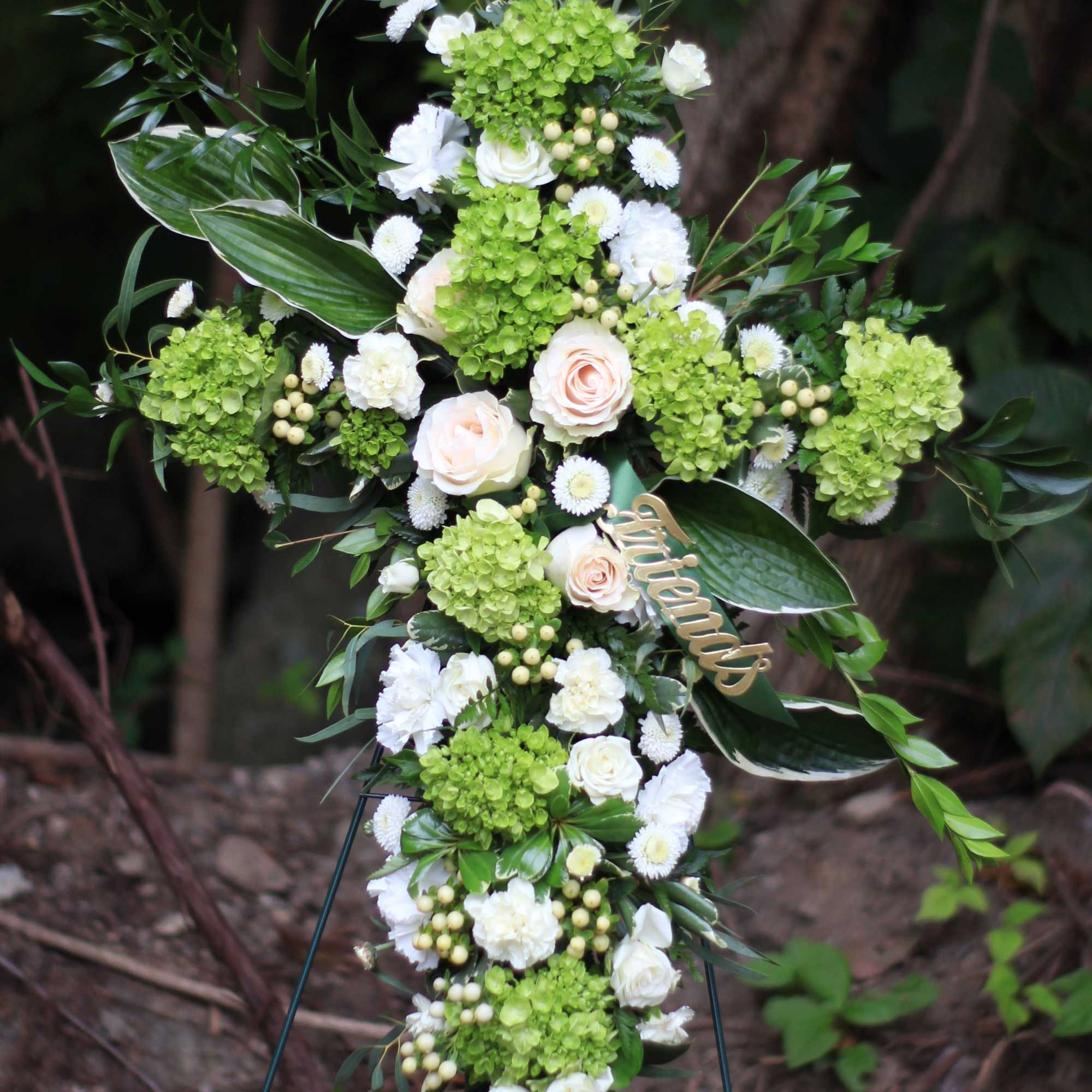 Cross-shaped floral arrangement with green hydrangeas, white roses, and a gold "friends" tag