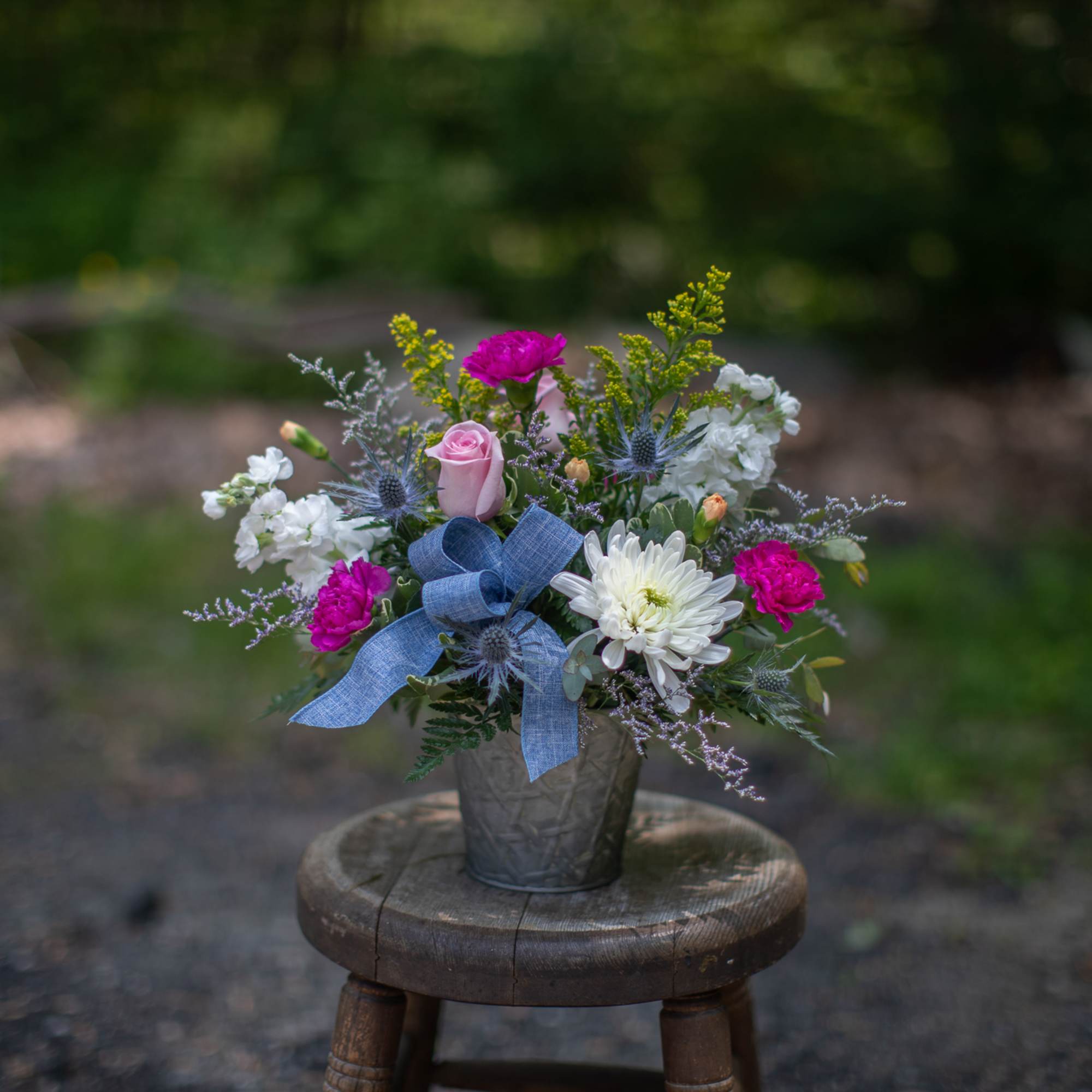 Mixed arrangement of pink and white flowers with a blue bow in a metal pot on a wooden stool outdoors