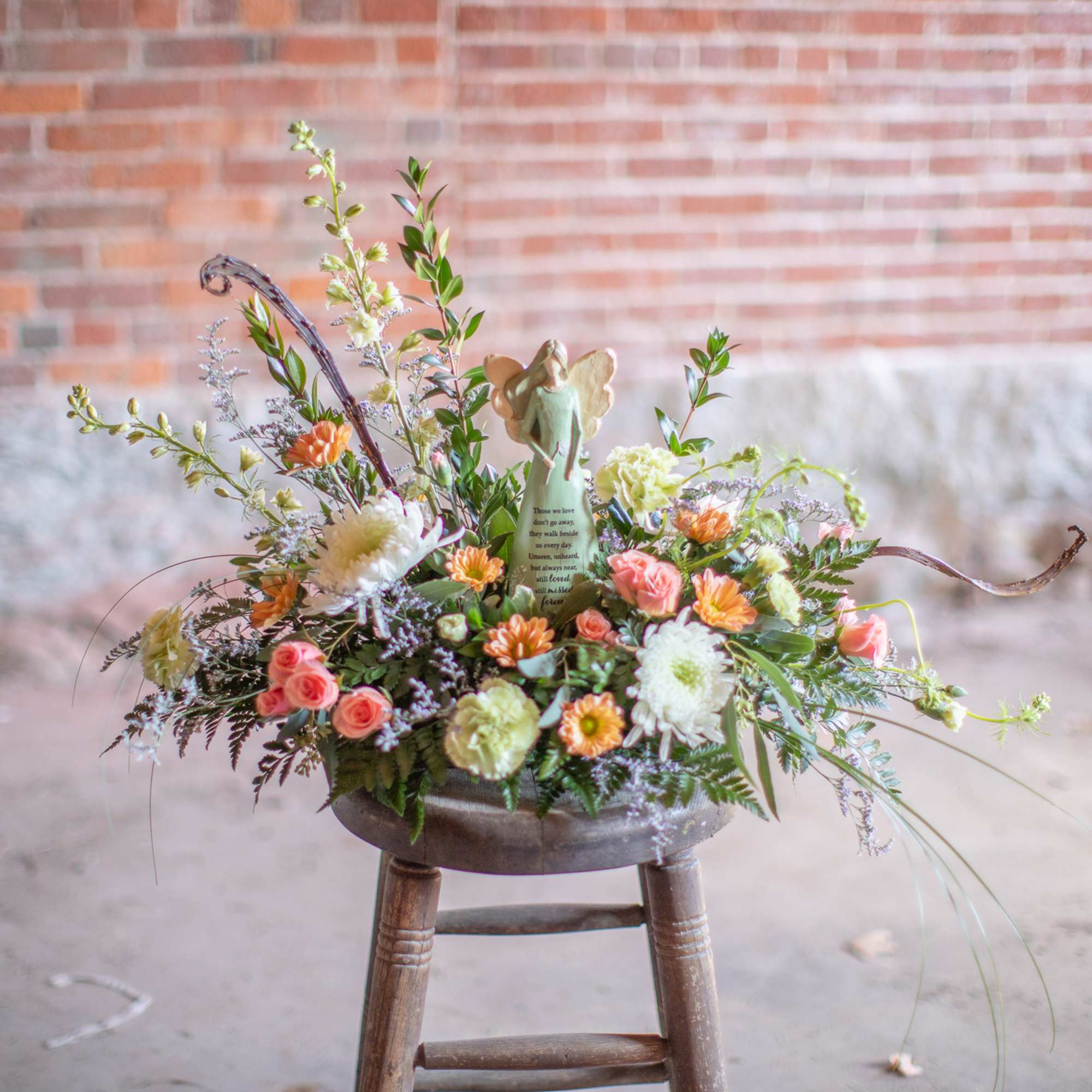 Low peach and white flower arrangement with angel figurine in a rustic bowl on a stool