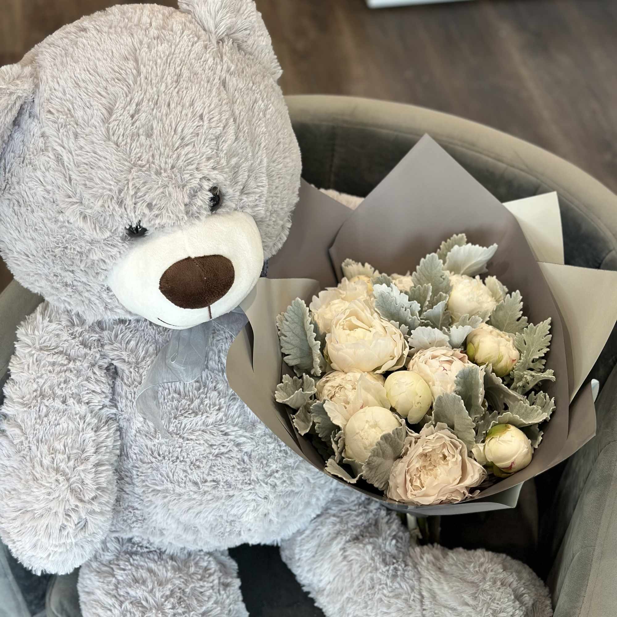A gray teddy bear sits beside a bouquet of pale cream flowers.