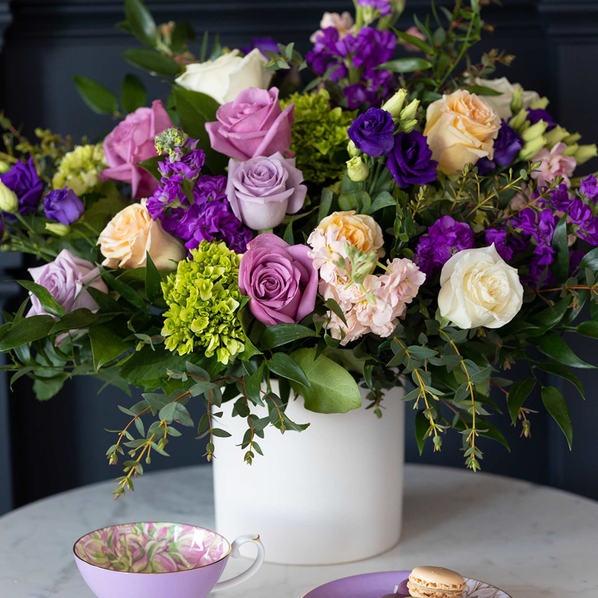 Mixed bouquet of roses and purple flowers in a white vase with teacup and macarons