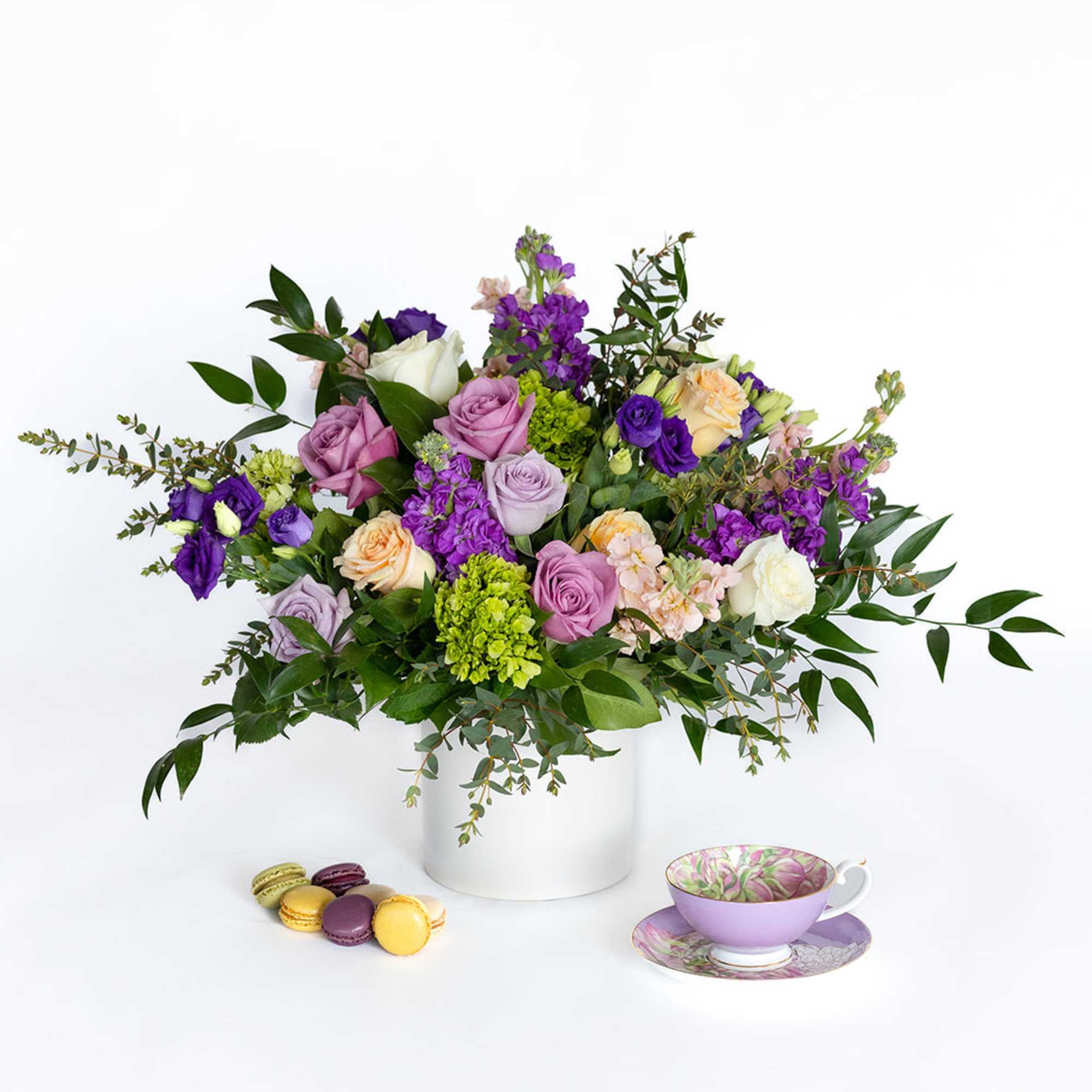 Mixed bouquet of purple, pink, white, and peach roses in a white vase with macarons and a teacup