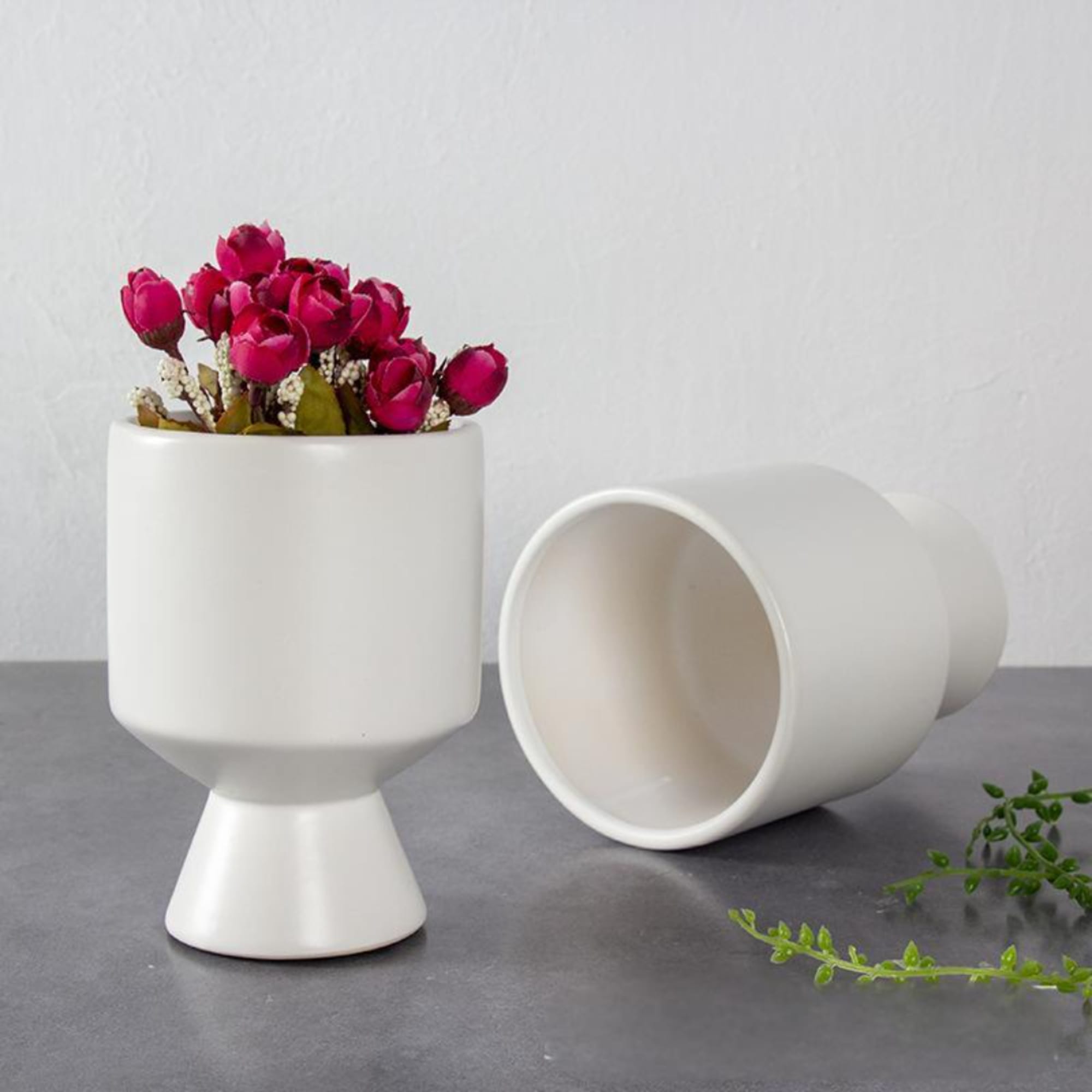 Pink flowers in a white pedestal vase beside a matching vase on its side
