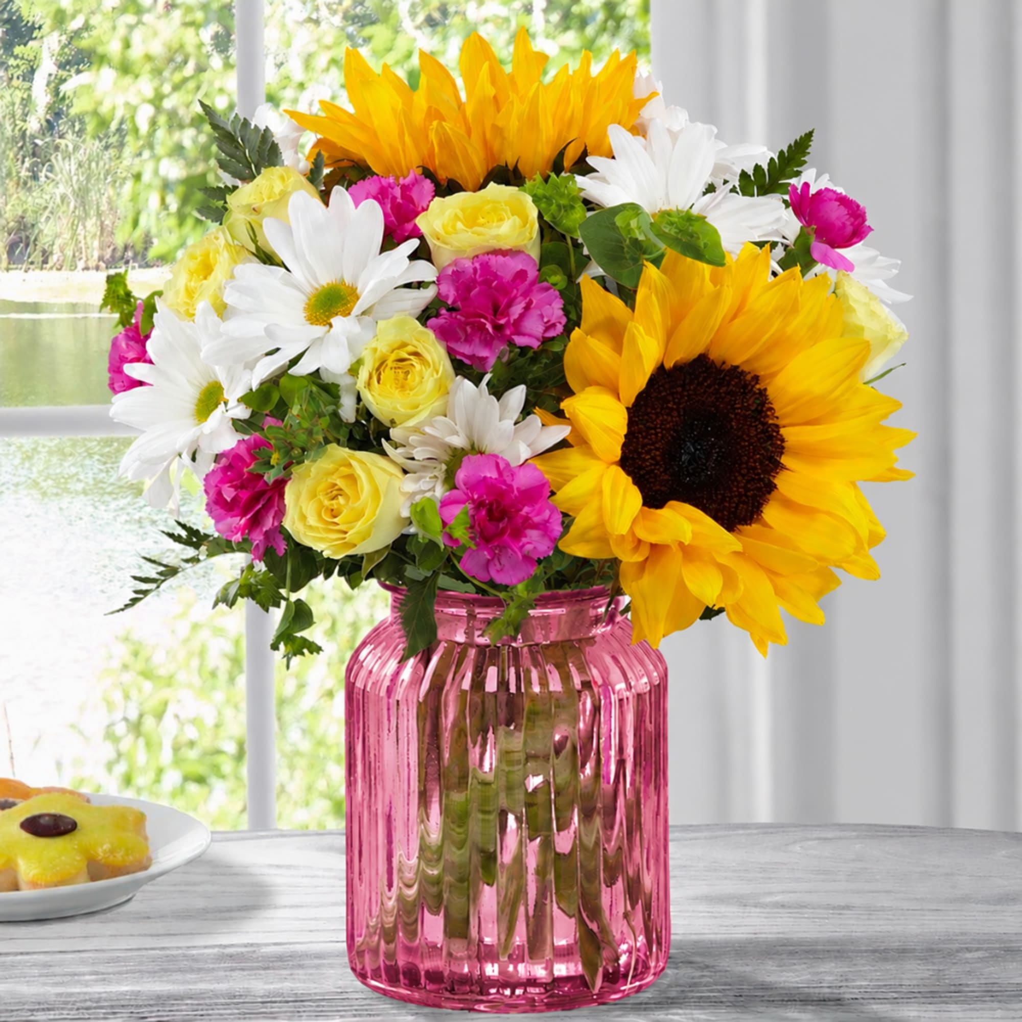 Bouquet of sunflowers, white daisies, pink carnations, and yellow roses in a pink glass vase