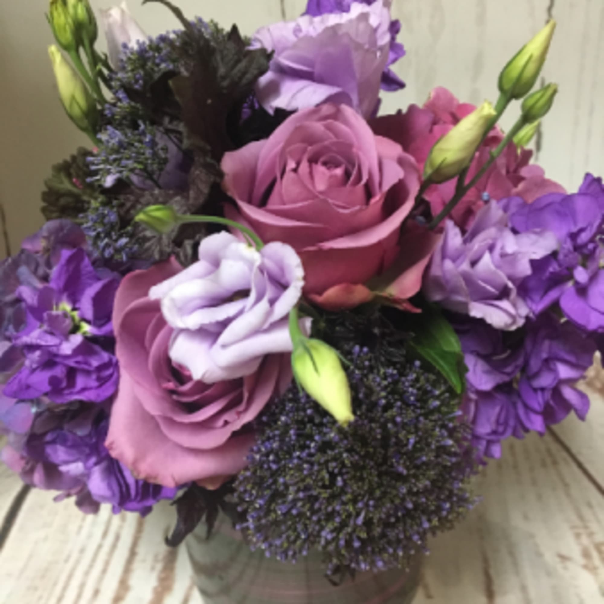 Compact arrangement of purple and lavender roses and blooms in a short clear glass vase