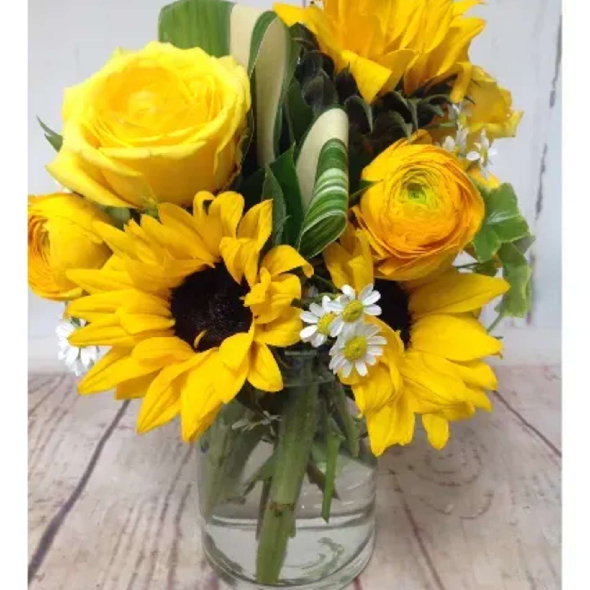 Yellow sunflowers, rose, and ranunculus arranged in a clear glass jar on a wooden surface