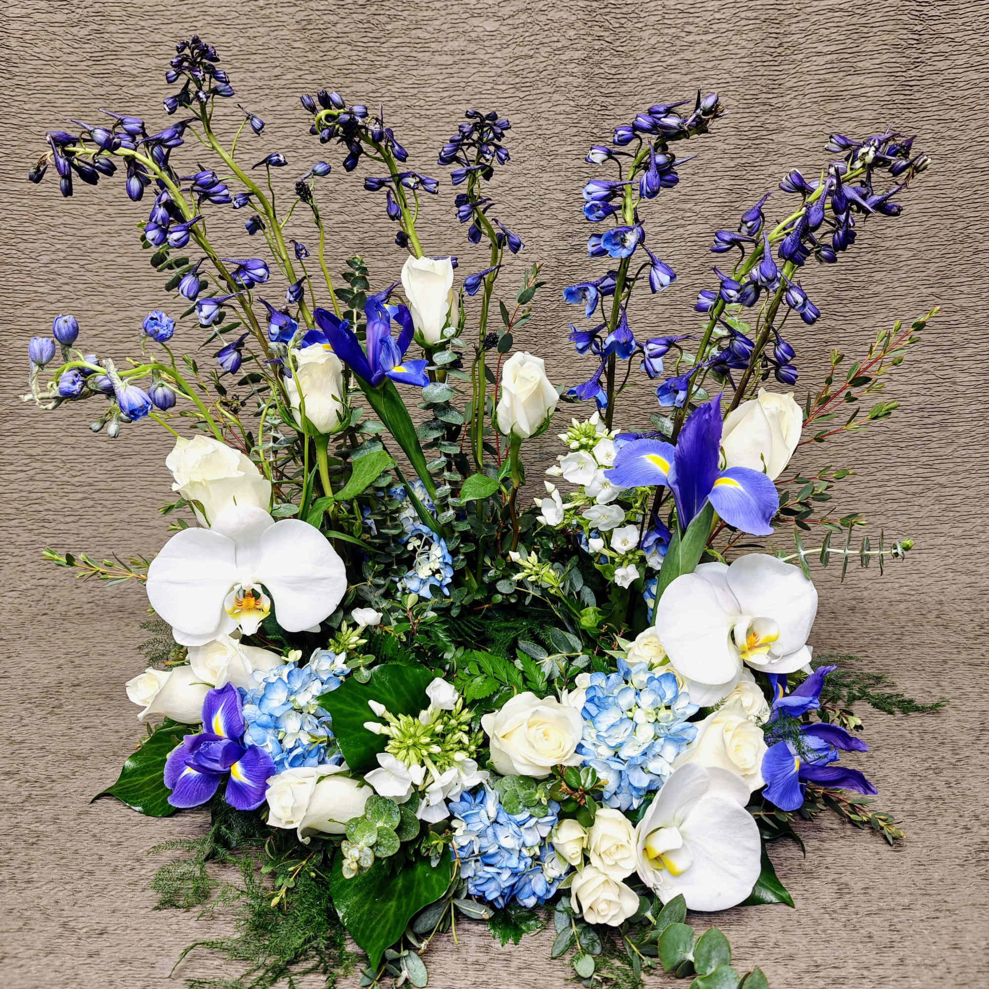Low arrangement of white orchids, white roses, and blue hydrangeas with tall blue blooms.