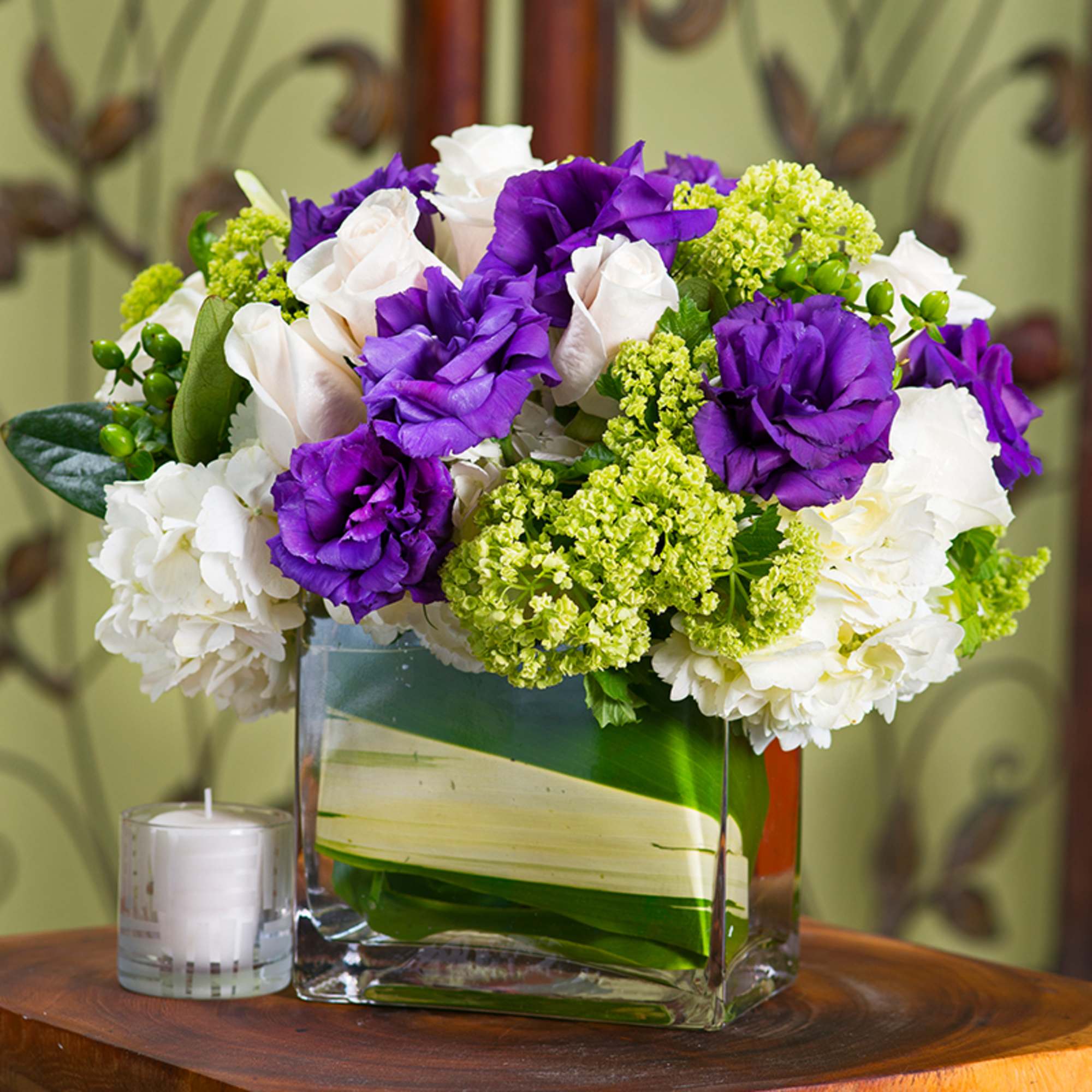 Purple and white bouquet in a square glass vase with a candle beside it