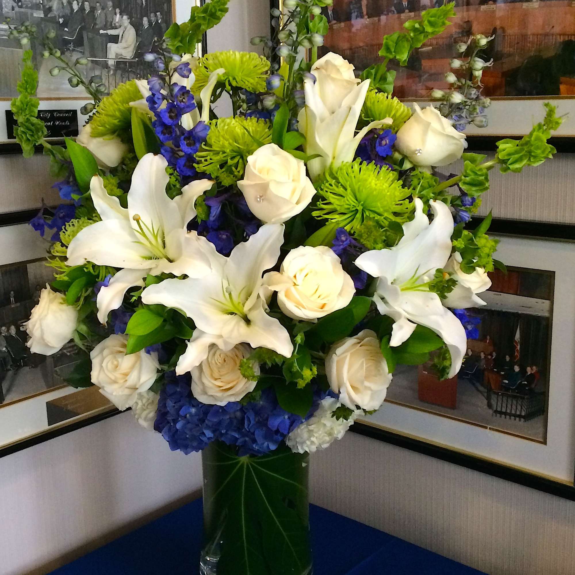 White lilies and roses with blue and green flowers in a tall glass vase