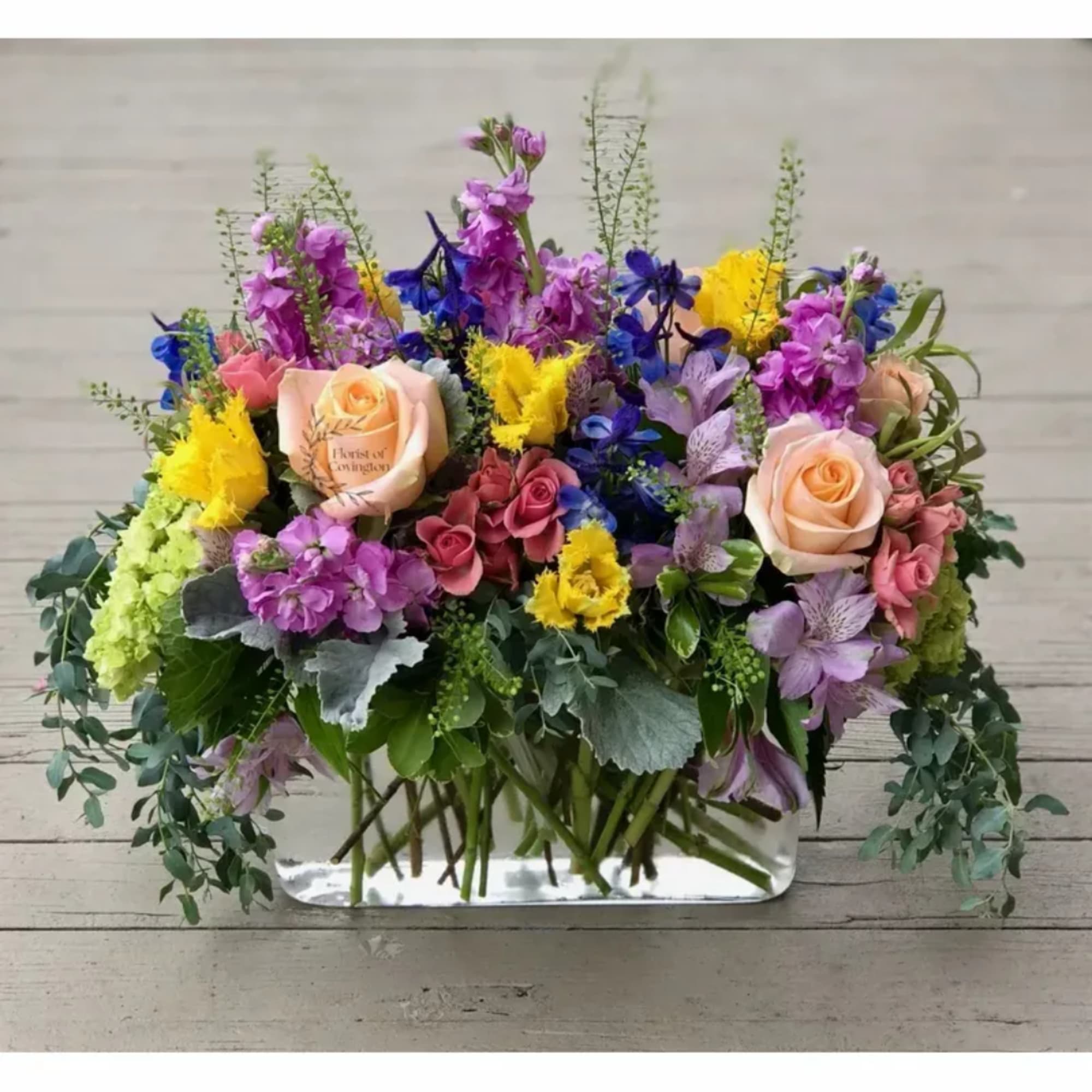 Mixed bouquet of roses, purple blooms, and yellow flowers in a low rectangular vase
