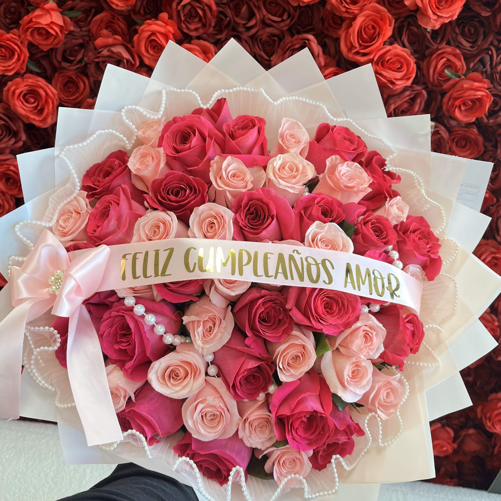 Round bouquet of hot pink and light pink roses with white wrap, pearls, and a Spanish birthday ribbon.