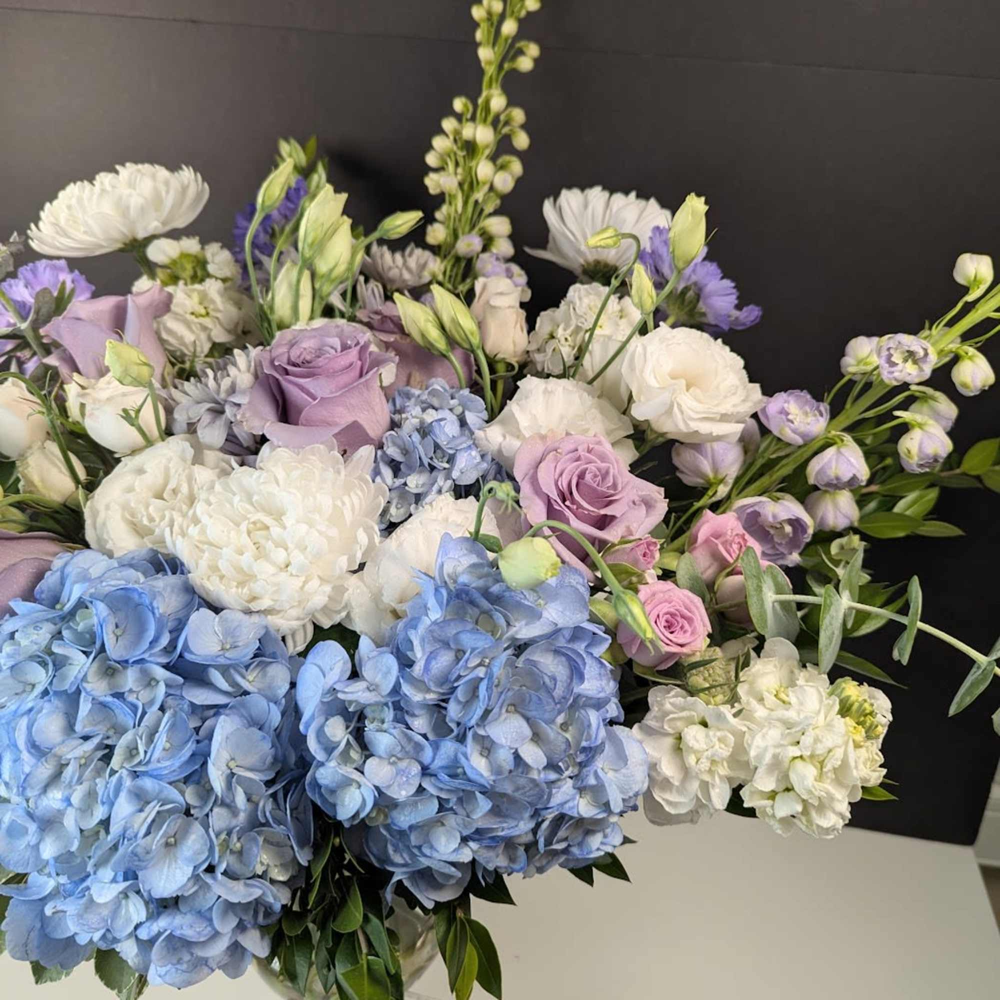 Pastel bouquet of blue hydrangeas, lavender roses, and white blooms in a clear vase