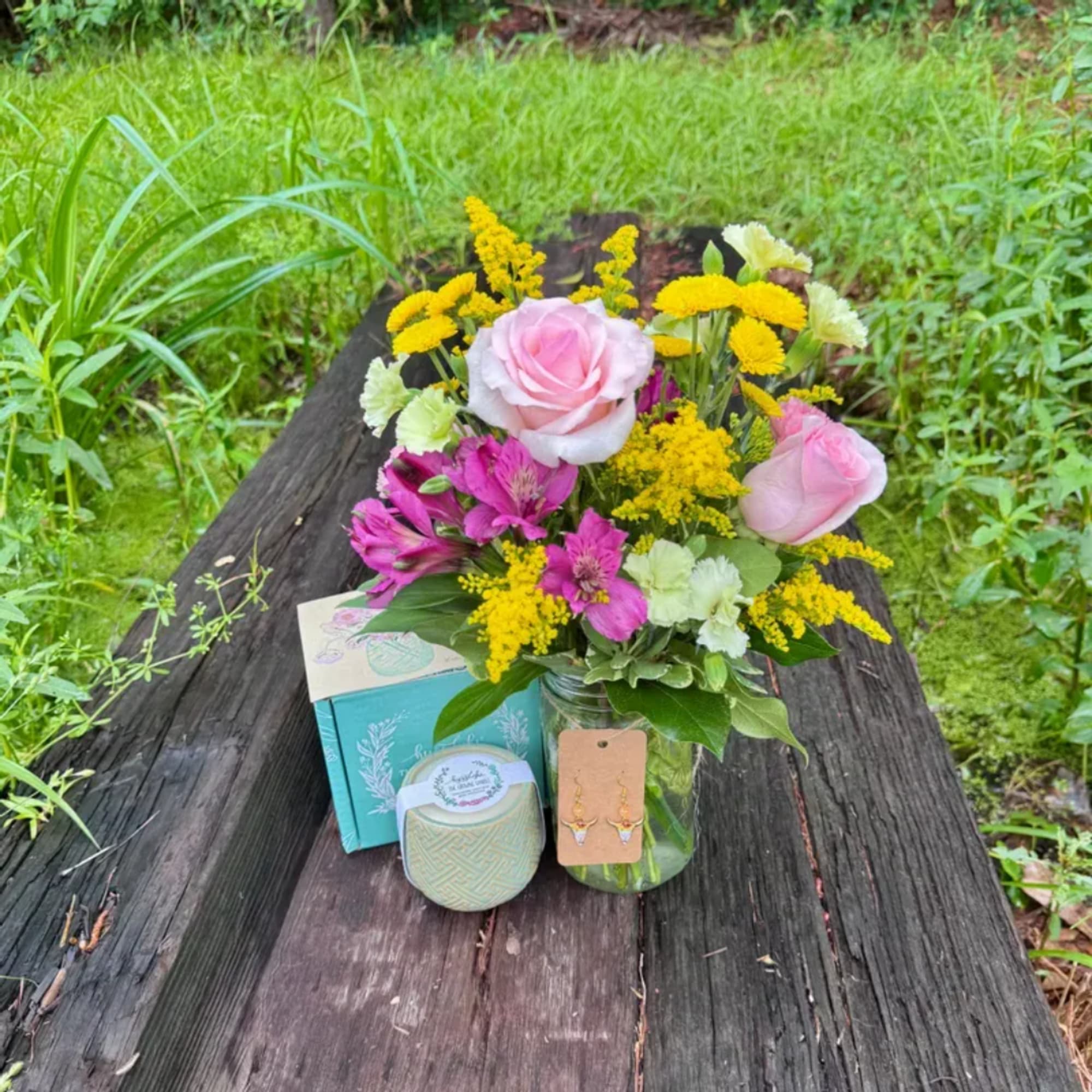 Pink roses and yellow flowers arranged in a glass jar with small gift items beside it