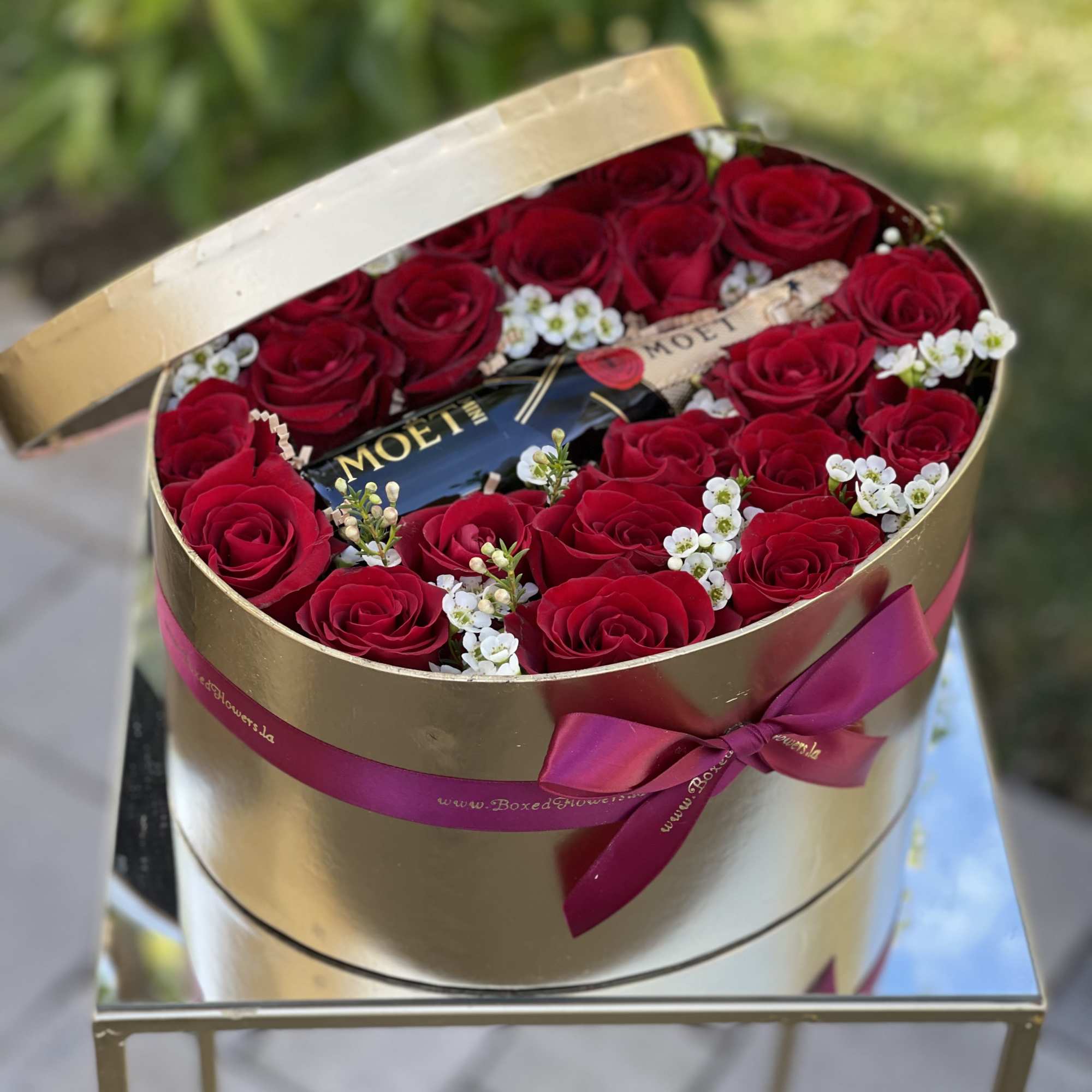 Red roses arranged in a gold round box with a champagne bottle and ribbon