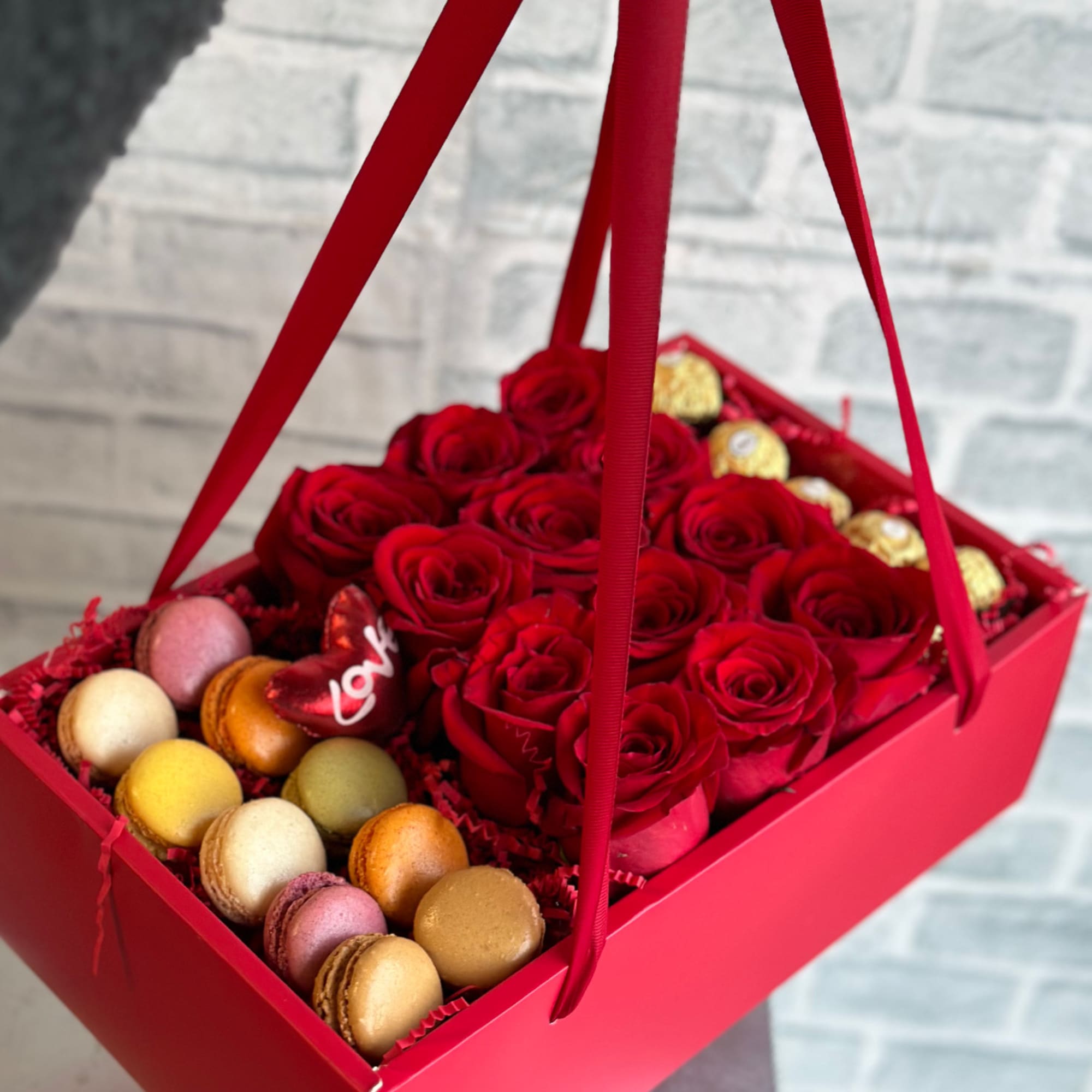 Red roses and assorted macarons in a red gift box with ribbon handles