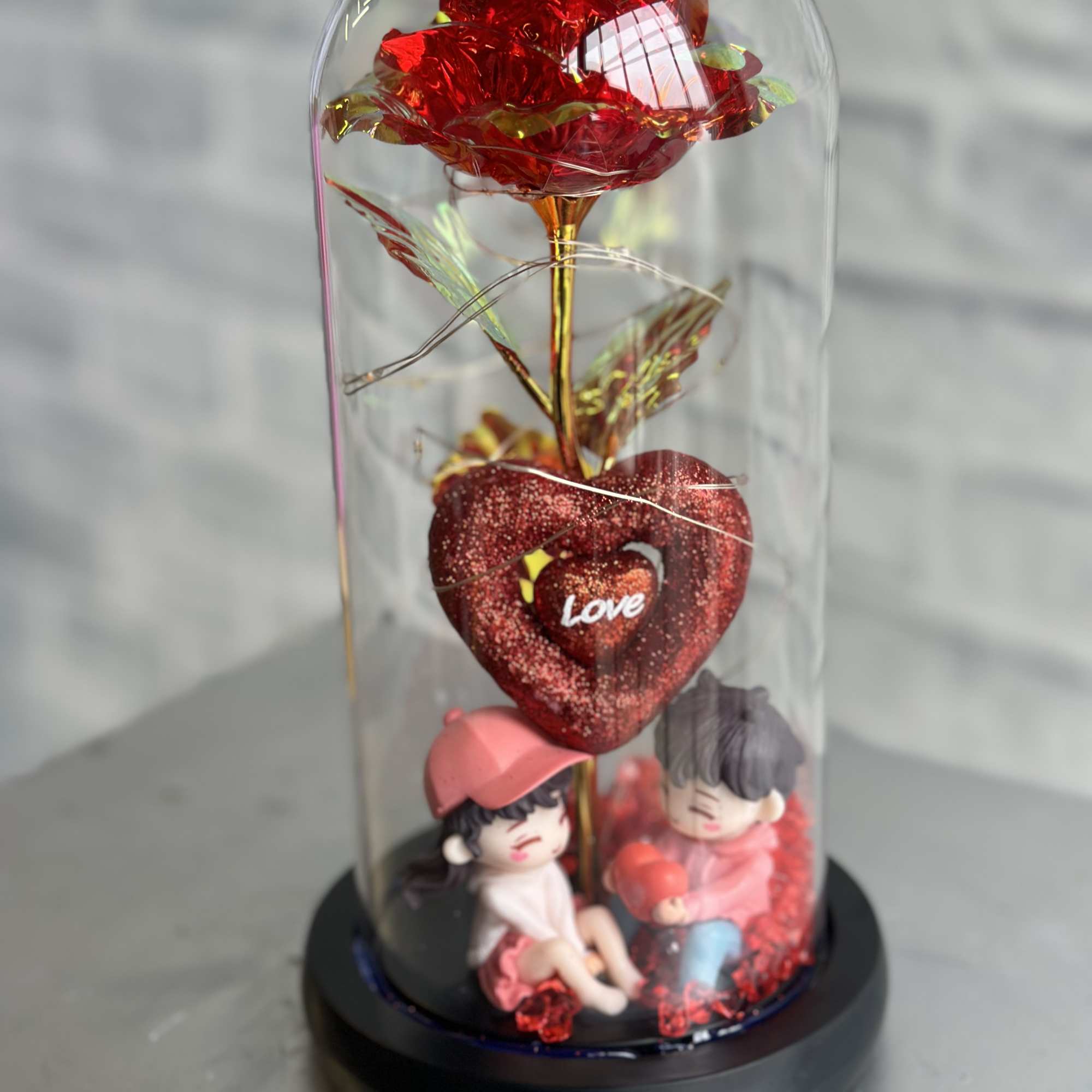 Red decorative rose under a clear dome with two small figurines at the base