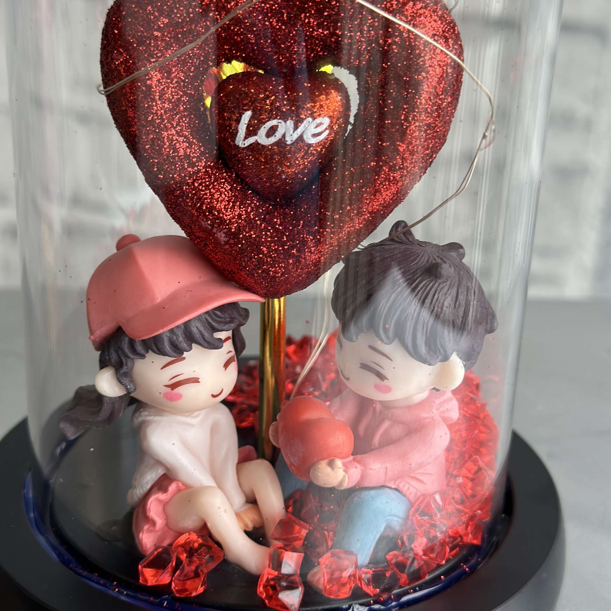 Two figurines under a glittery red heart in a glass dome