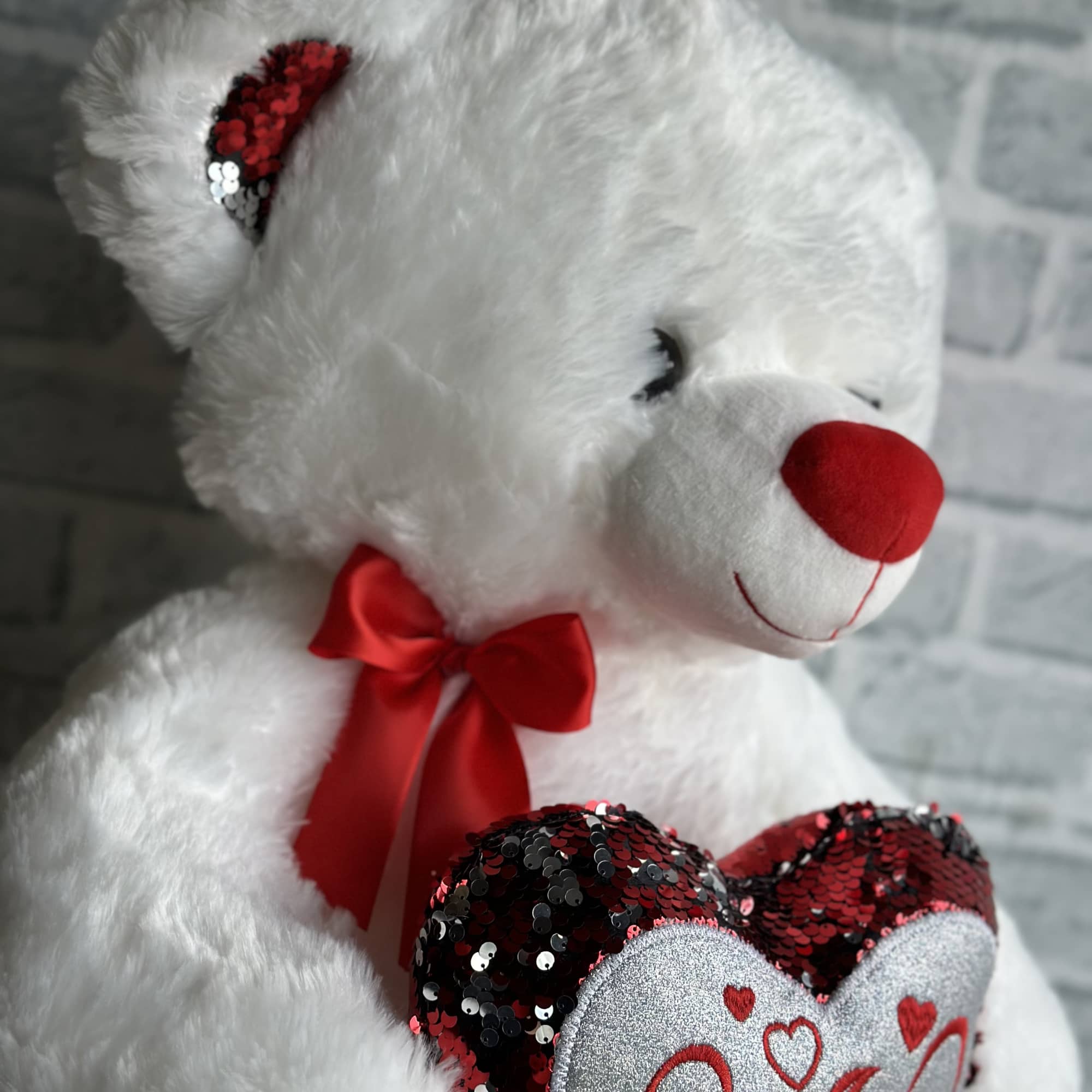 White teddy bear holding a sequined heart with a red bow