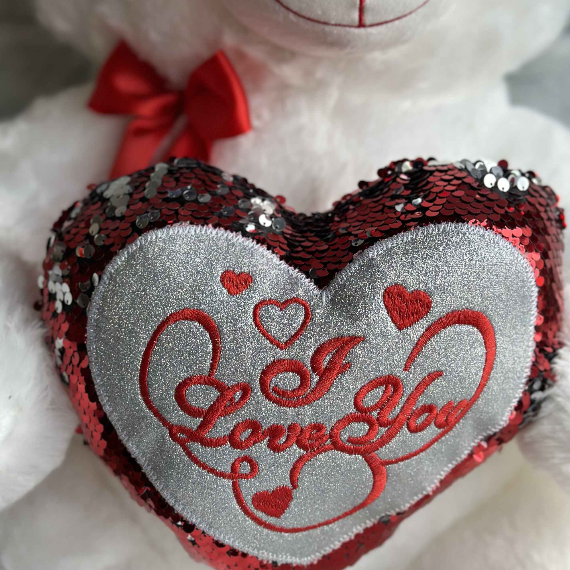 White teddy bear holding a sequined heart pillow with "I Love You" text