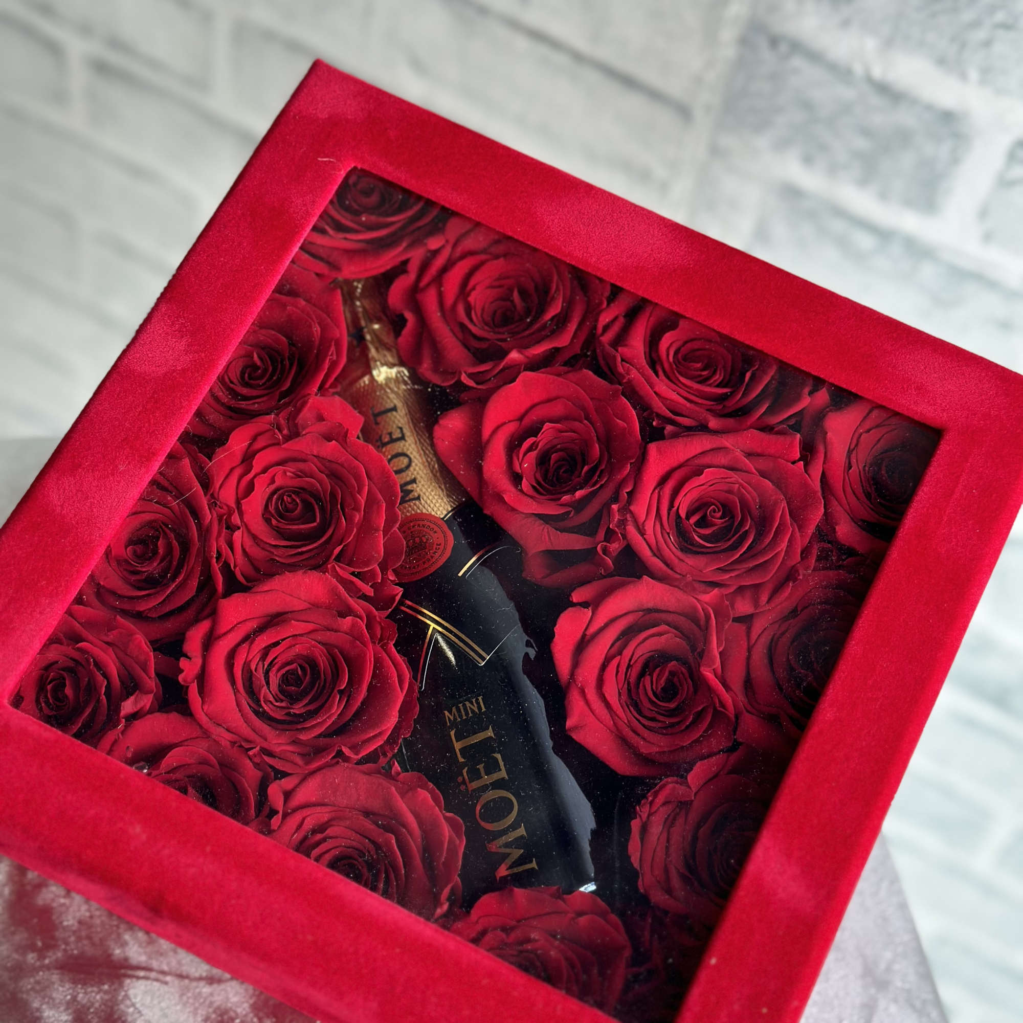 Red roses arranged in a square gift box with a bottle in the center