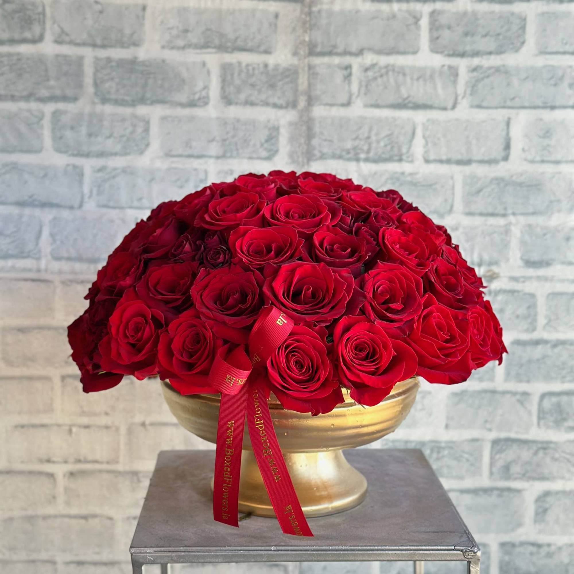Round arrangement of red roses in a gold pedestal bowl with a red ribbon