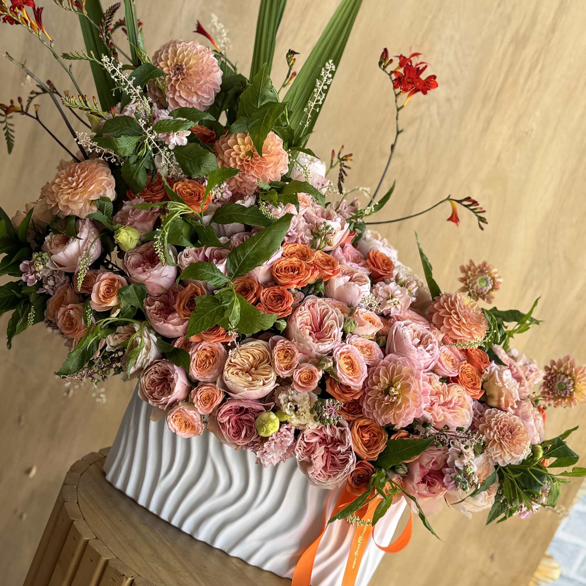Large pink and peach floral arrangement in a white vase with orange ribbon