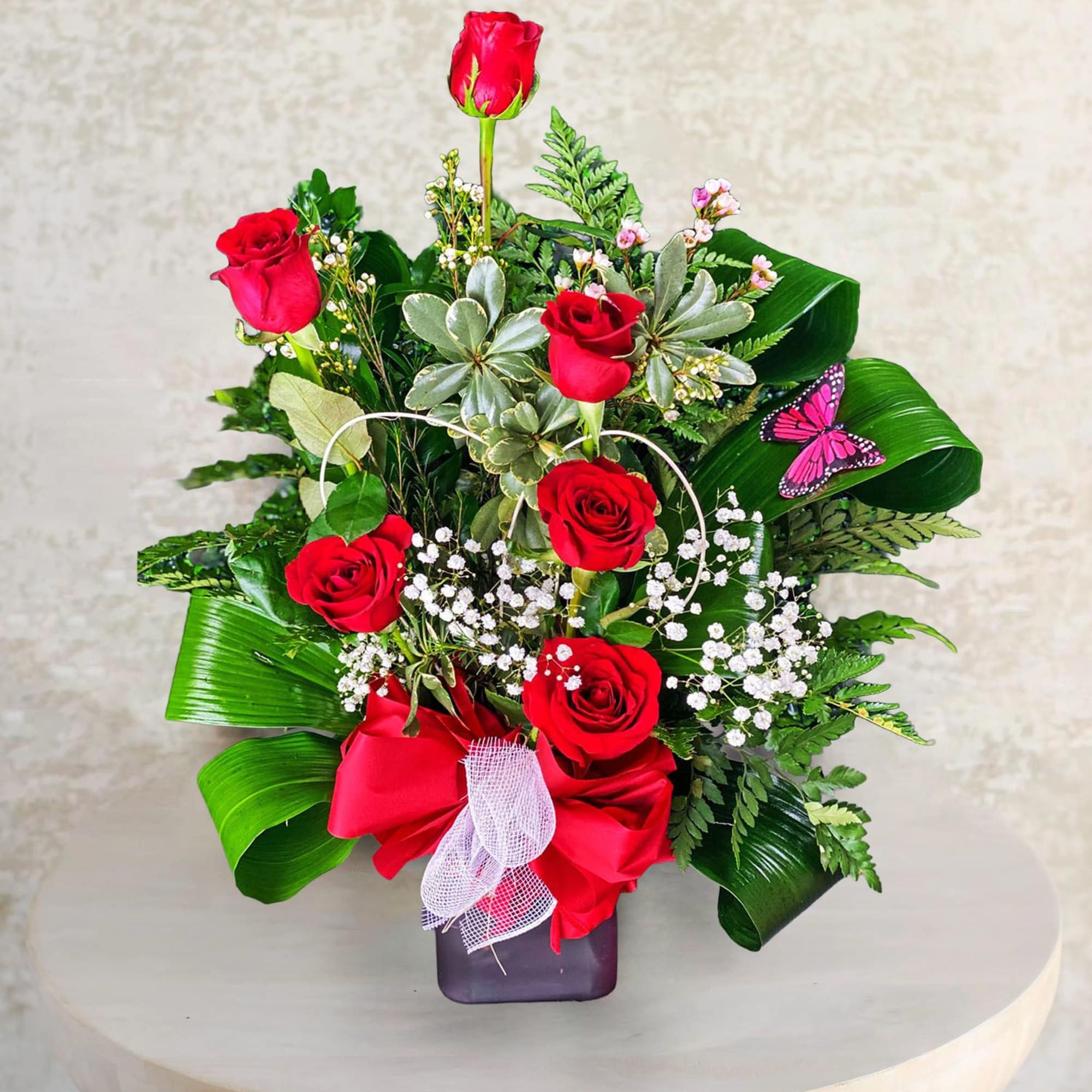 Red roses arranged in a square vase with greenery and a pink butterfly accent
