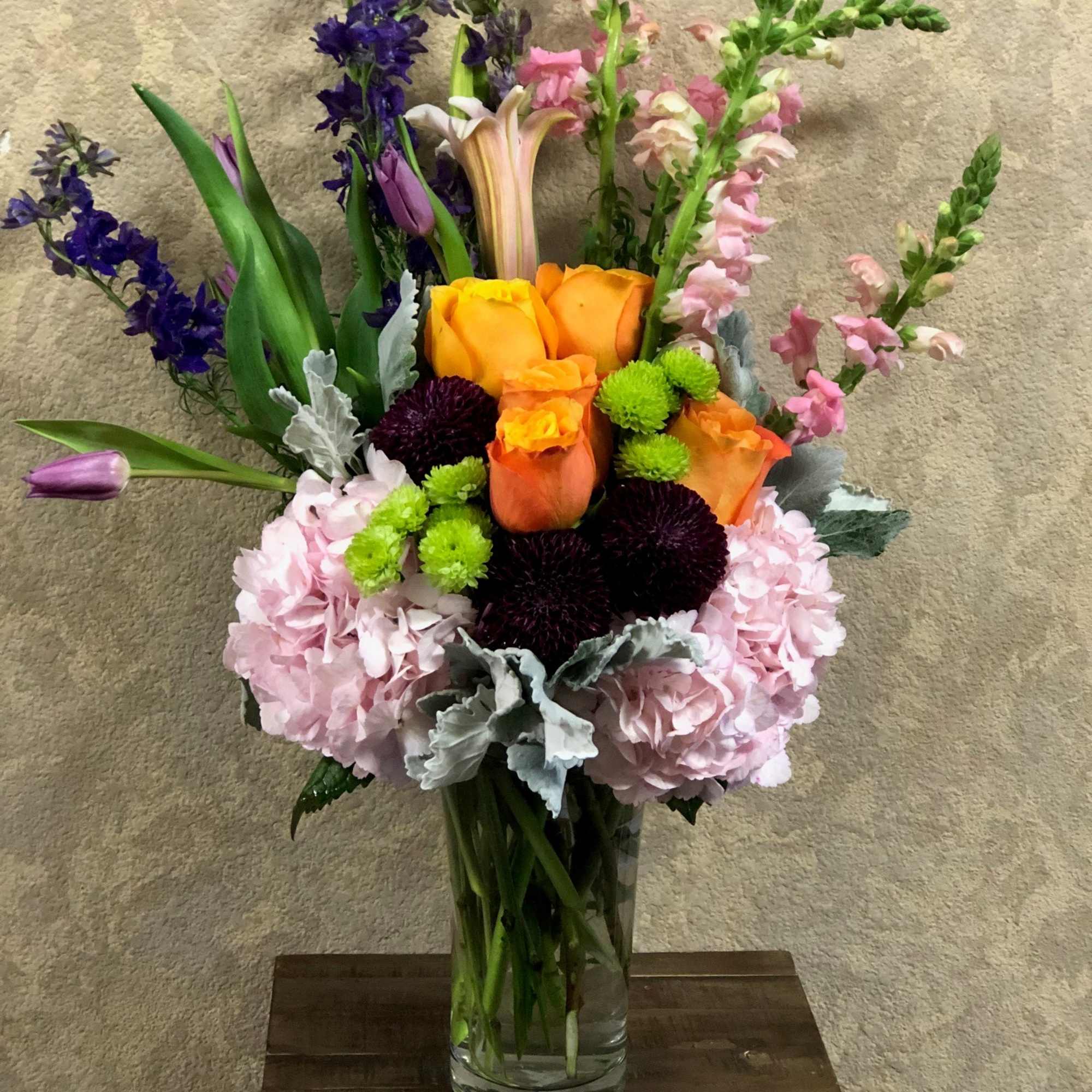 Mixed bouquet of orange roses, pink hydrangeas, and purple flowers in a glass vase