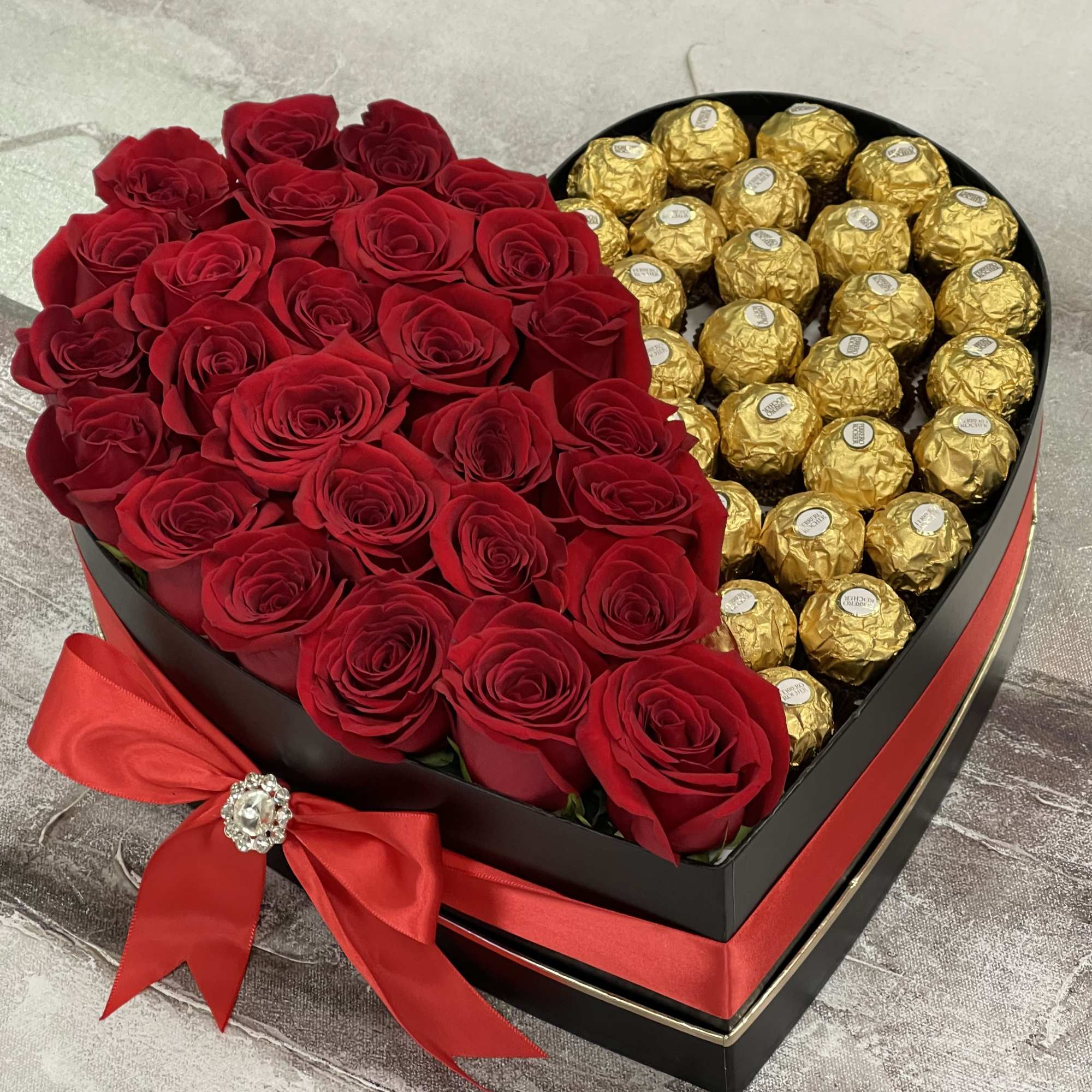 Heart-shaped black box filled with red roses and gold-wrapped chocolates tied with a red bow