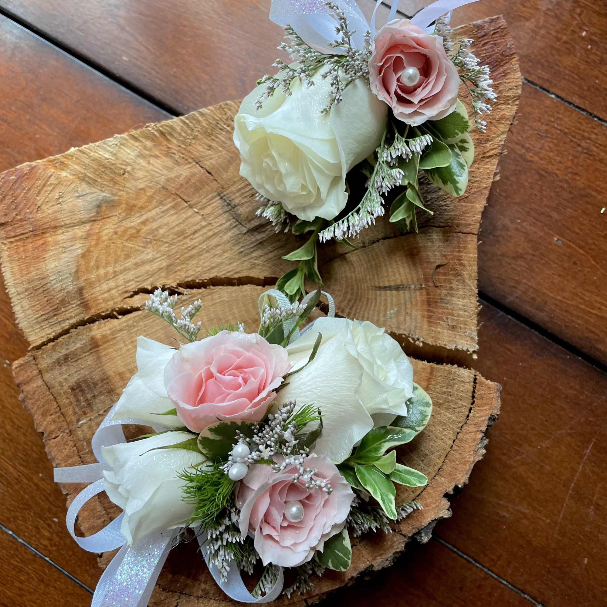 Two small corsages with white and pink roses, ribbon, and pearls displayed on rustic wood slices.