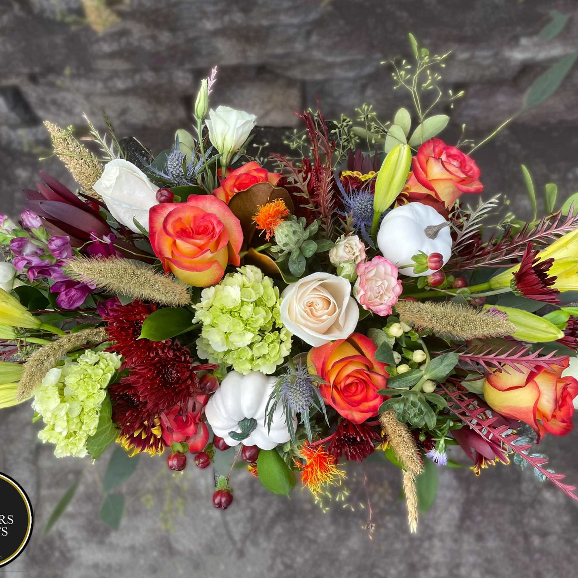Elongated fall flower arrangement with orange roses, yellow lilies, green hydrangeas, and white pumpkins.