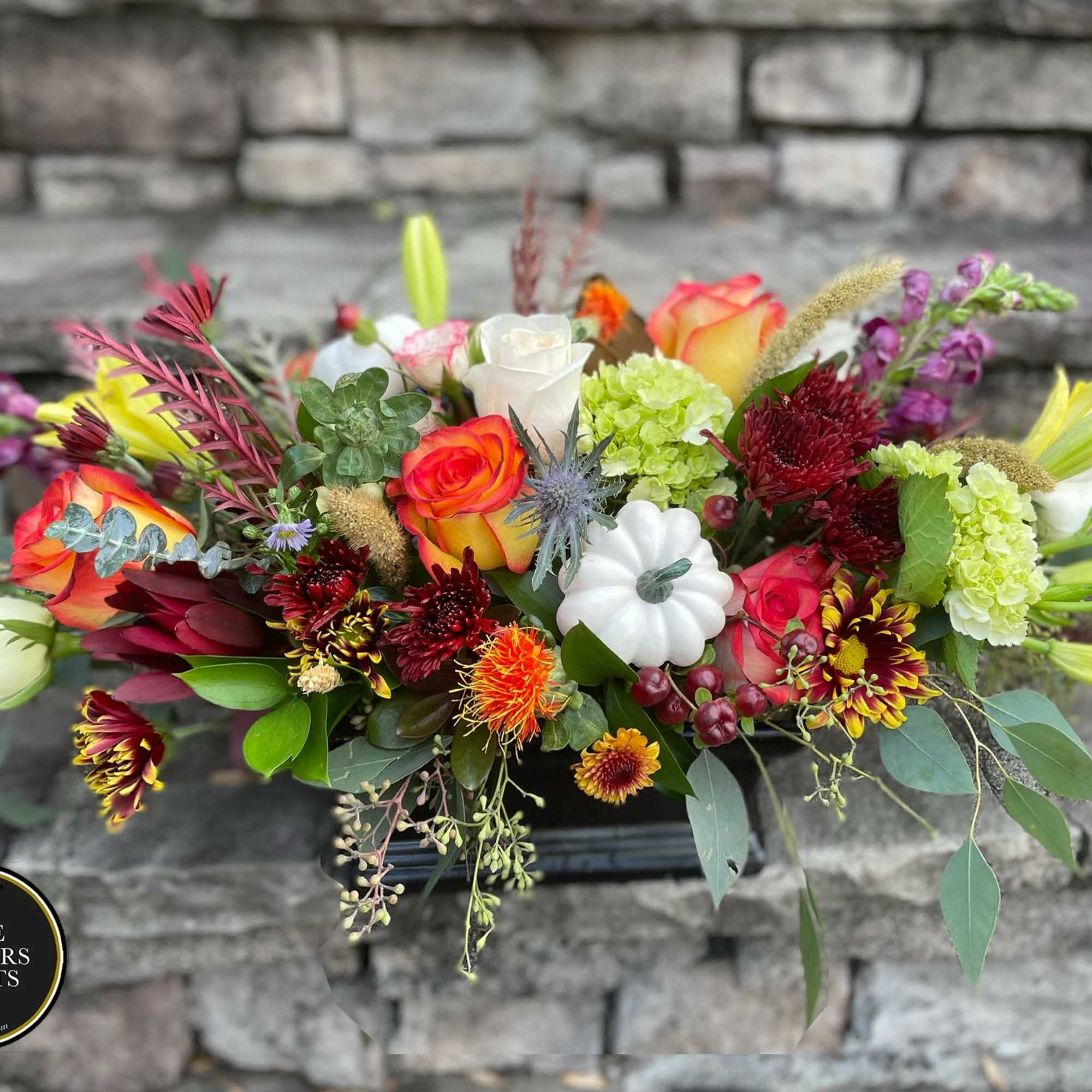 Low fall centerpiece with orange roses, mums, lilies, green hydrangea and a white mini pumpkin in a black container