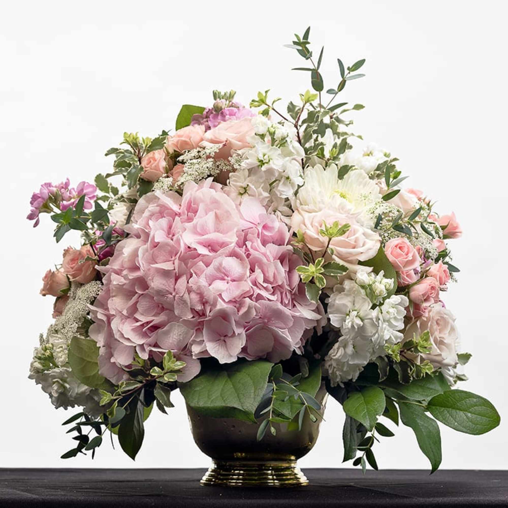 Low gold pedestal arrangement with pink hydrangea, pale roses, and white blooms.