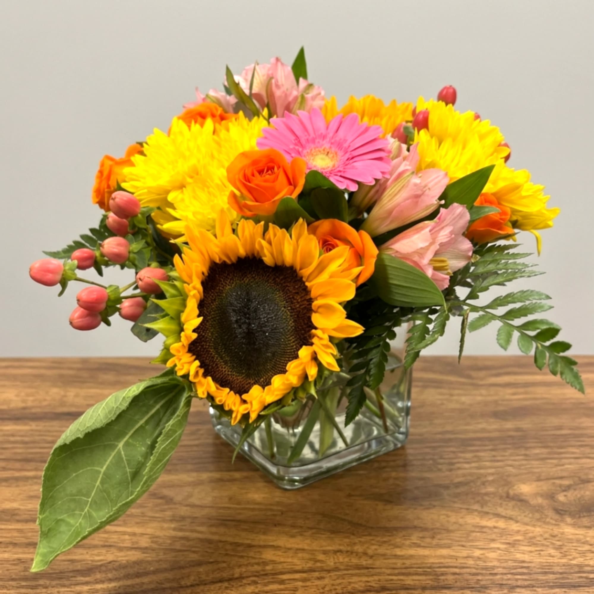 Bright mixed bouquet in a square glass vase with sunflowers, roses, and pink blooms