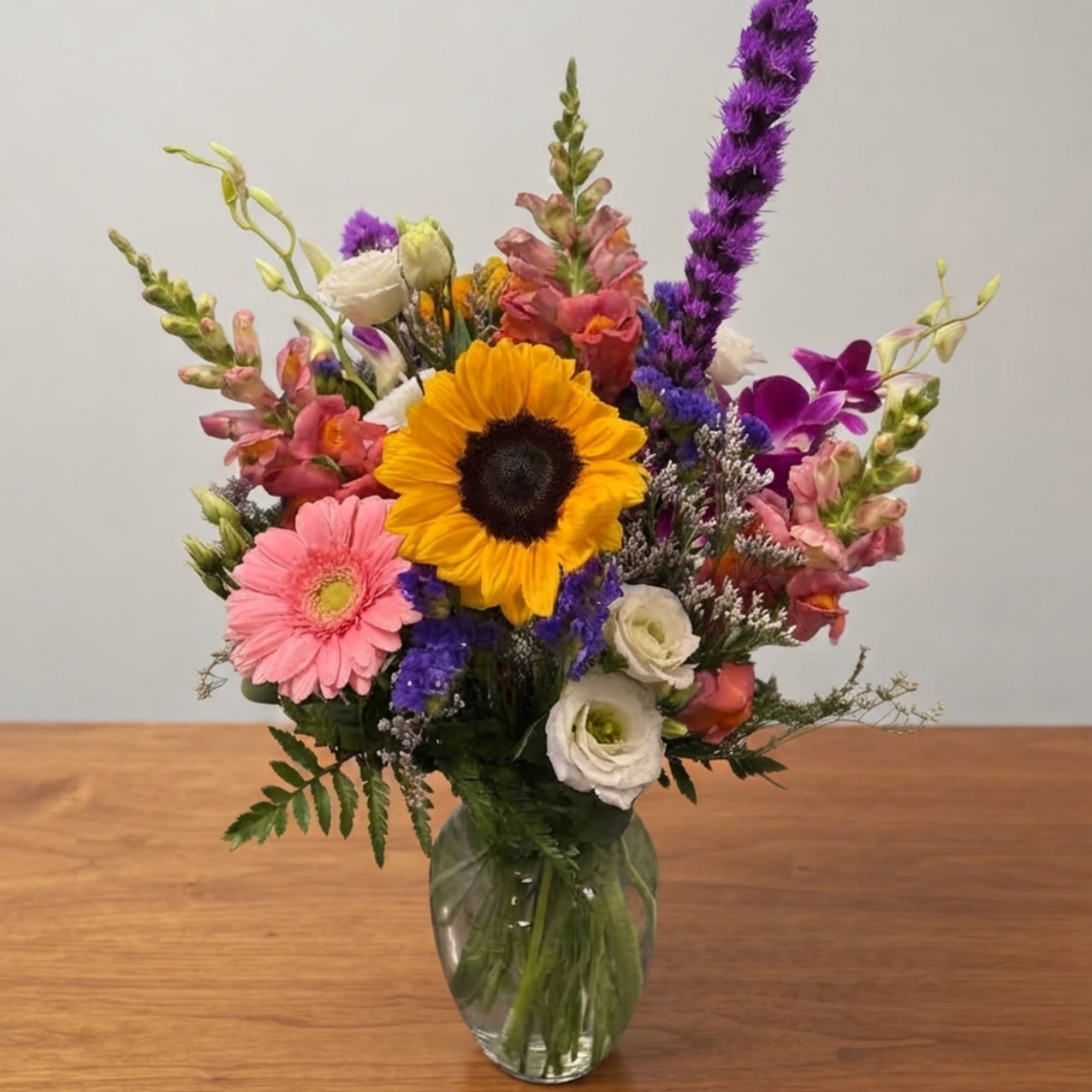 Mixed bouquet with a sunflower, pink gerbera, and purple blooms in a glass vase