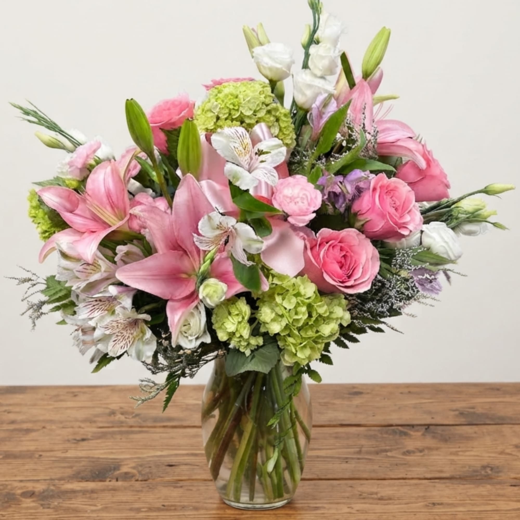 Pink lilies and roses arranged in a clear glass vase with green hydrangeas