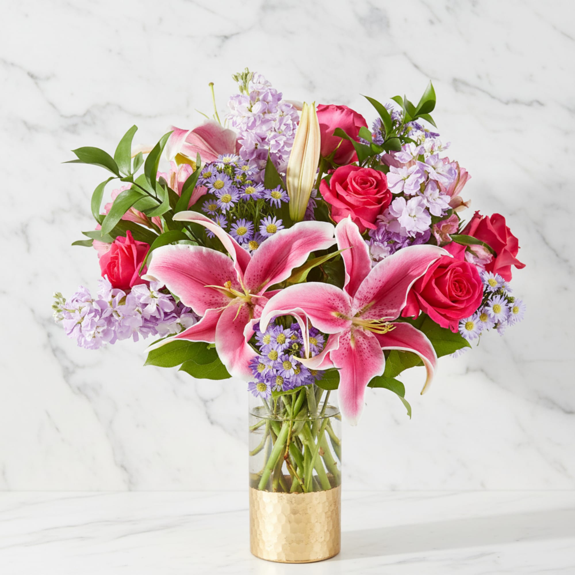 Pink lilies and roses arranged in a glass vase with purple filler flowers