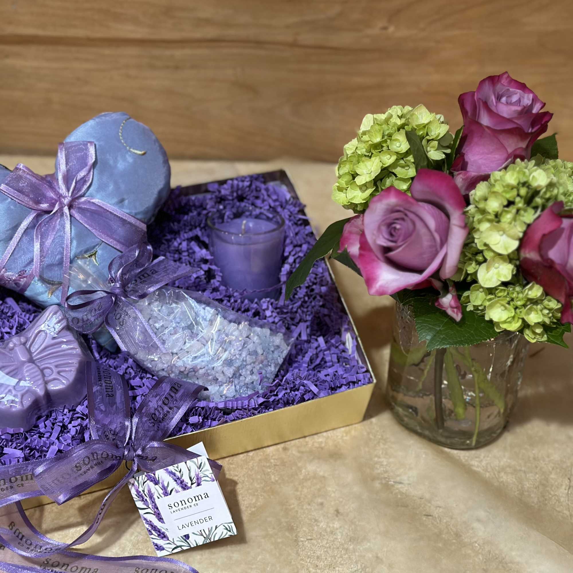 Pink roses and green hydrangeas in a glass vase beside a lavender gift box with candles and soap.