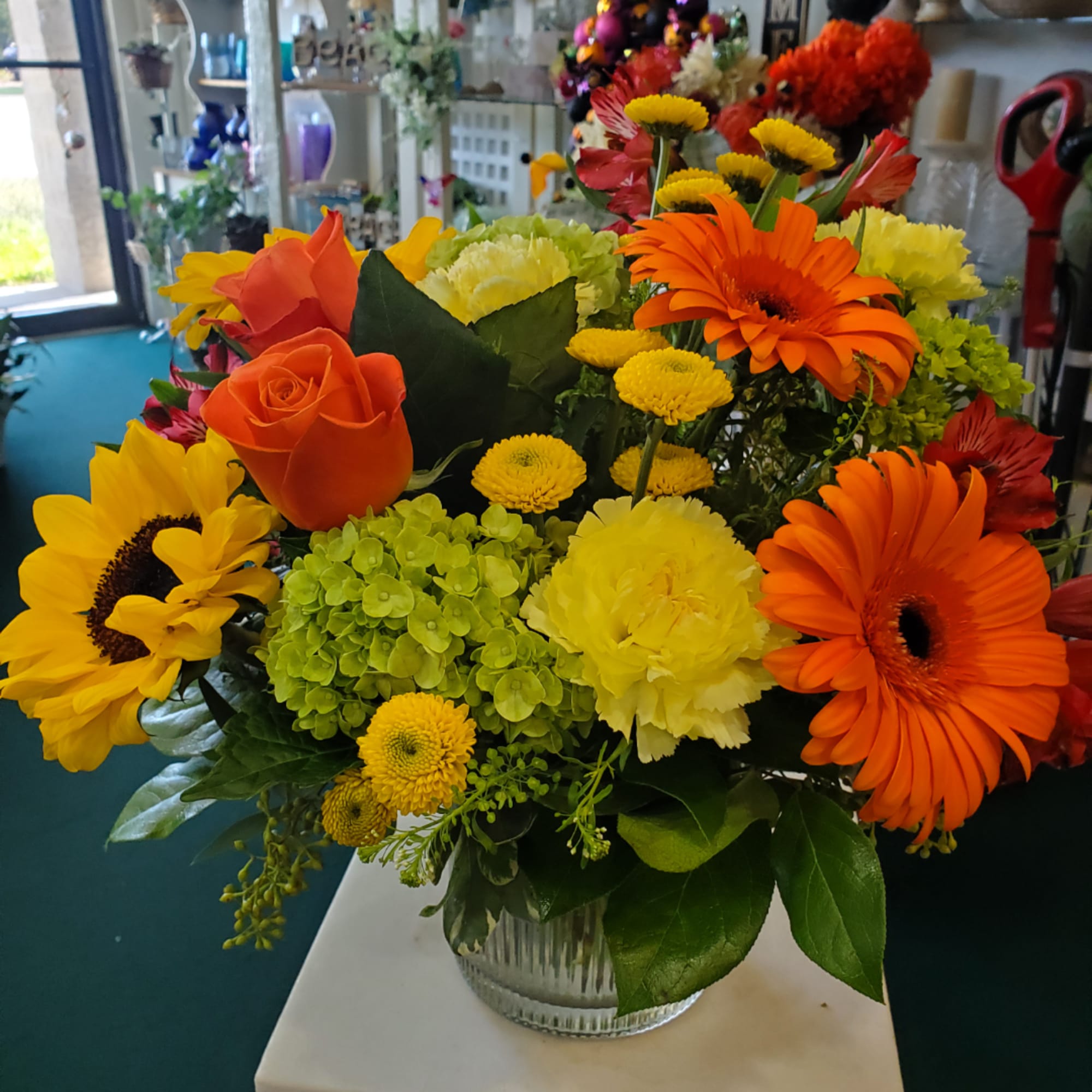 Bright mixed flower arrangement in a clear glass vase with sunflowers, roses, and gerbera daisies