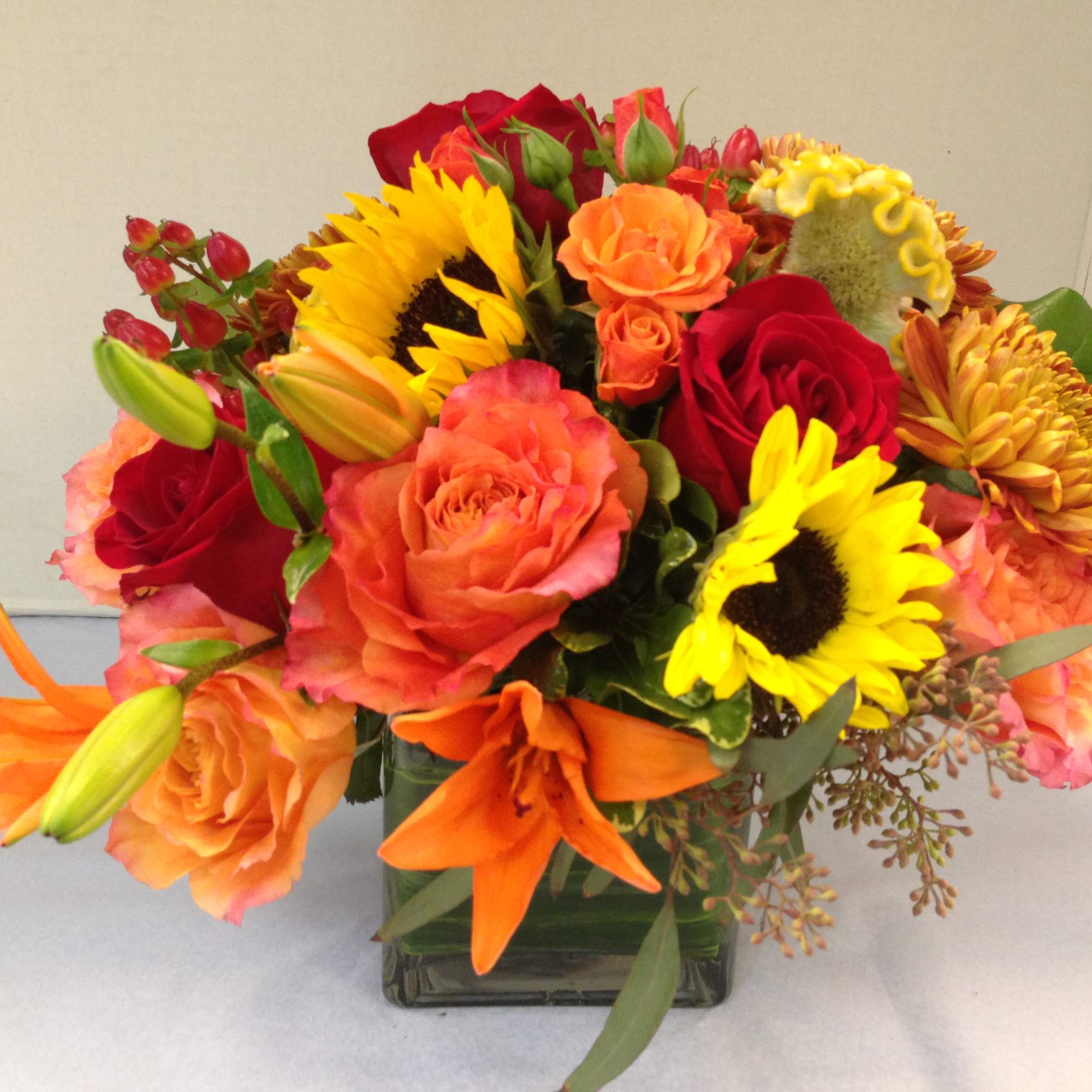 Bright mixed bouquet of roses, sunflowers, lilies, and mums in a square glass vase