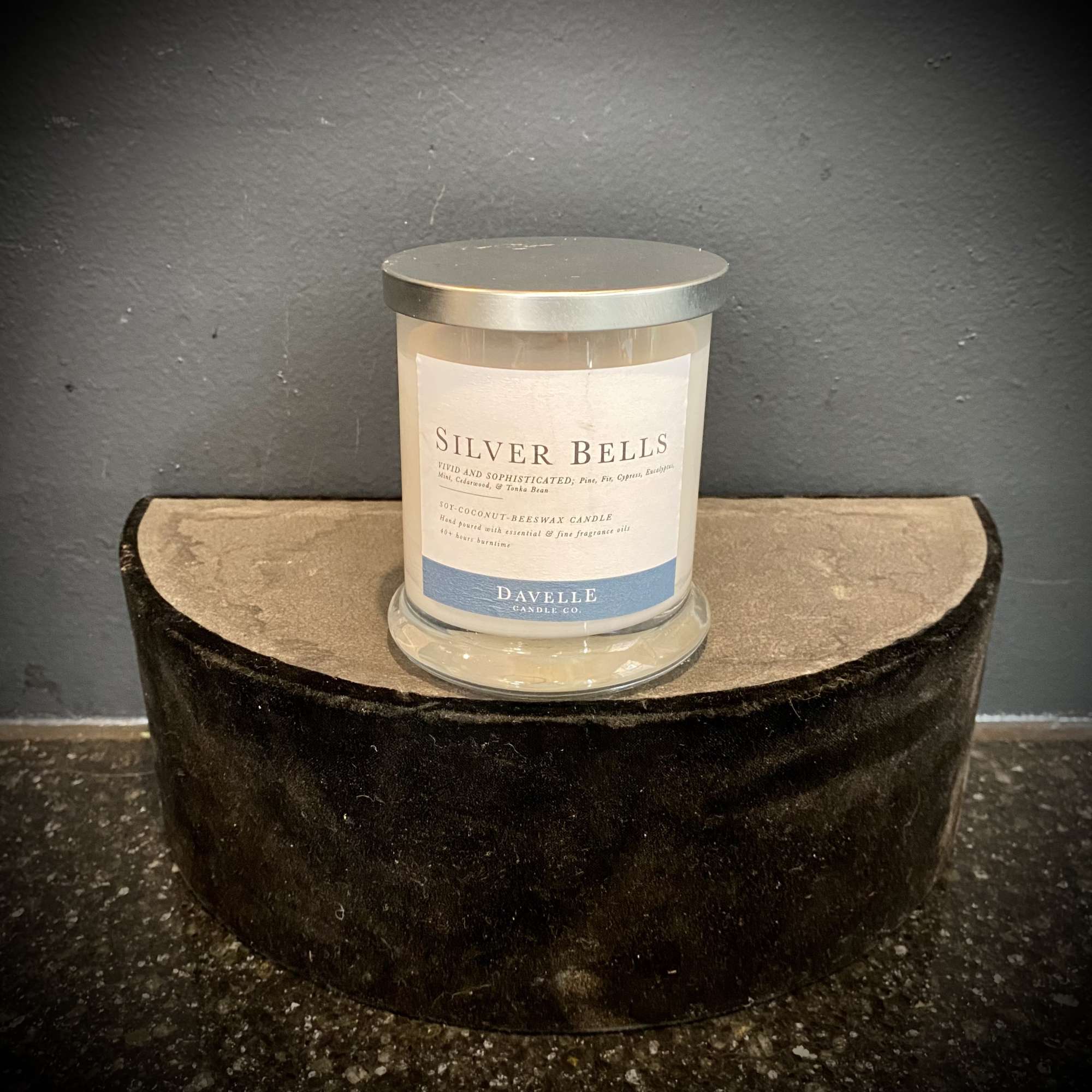 A scented candle in a glass jar with a silver lid sits on a velvet display stand.