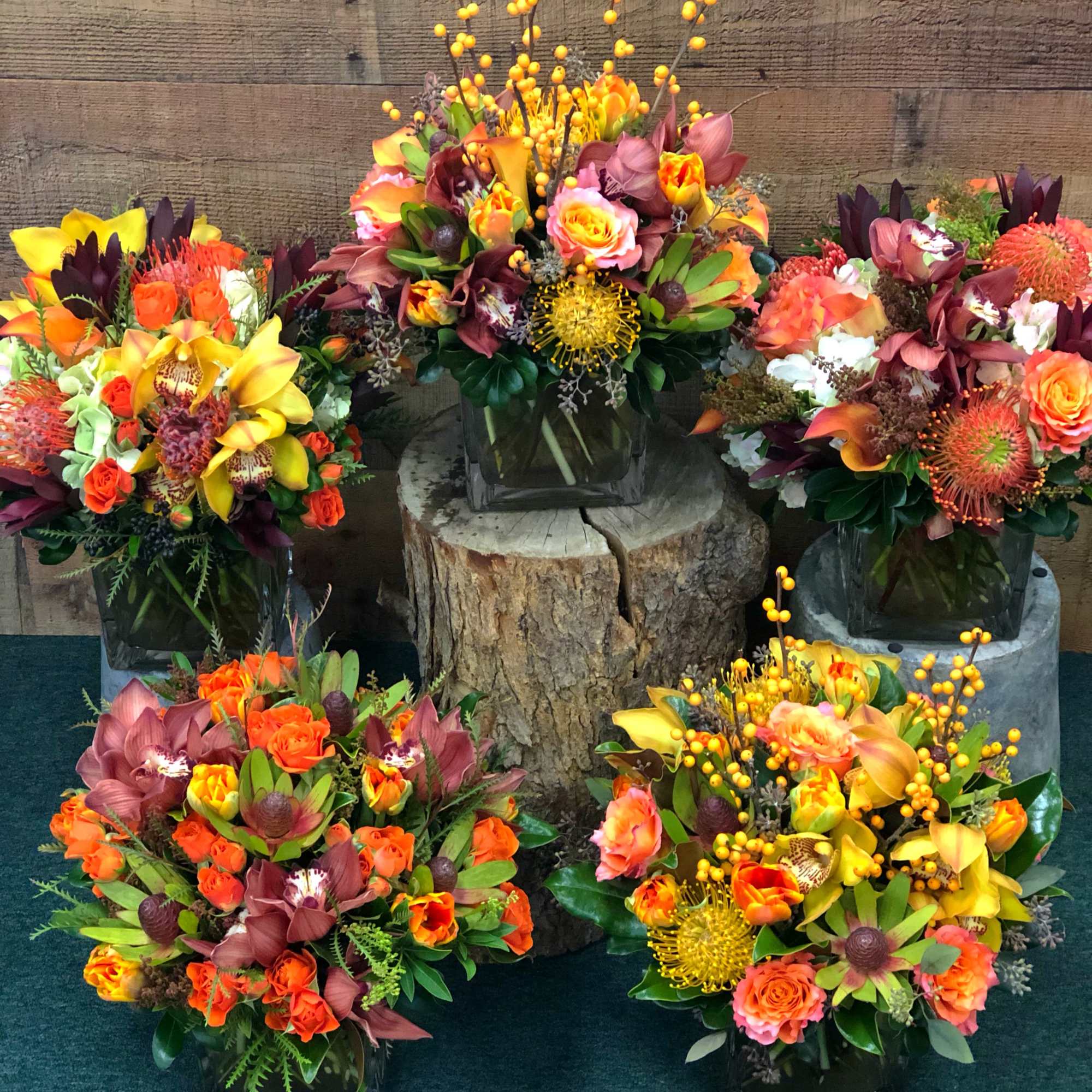 Five colorful floral arrangements in square glass vases around a tree stump