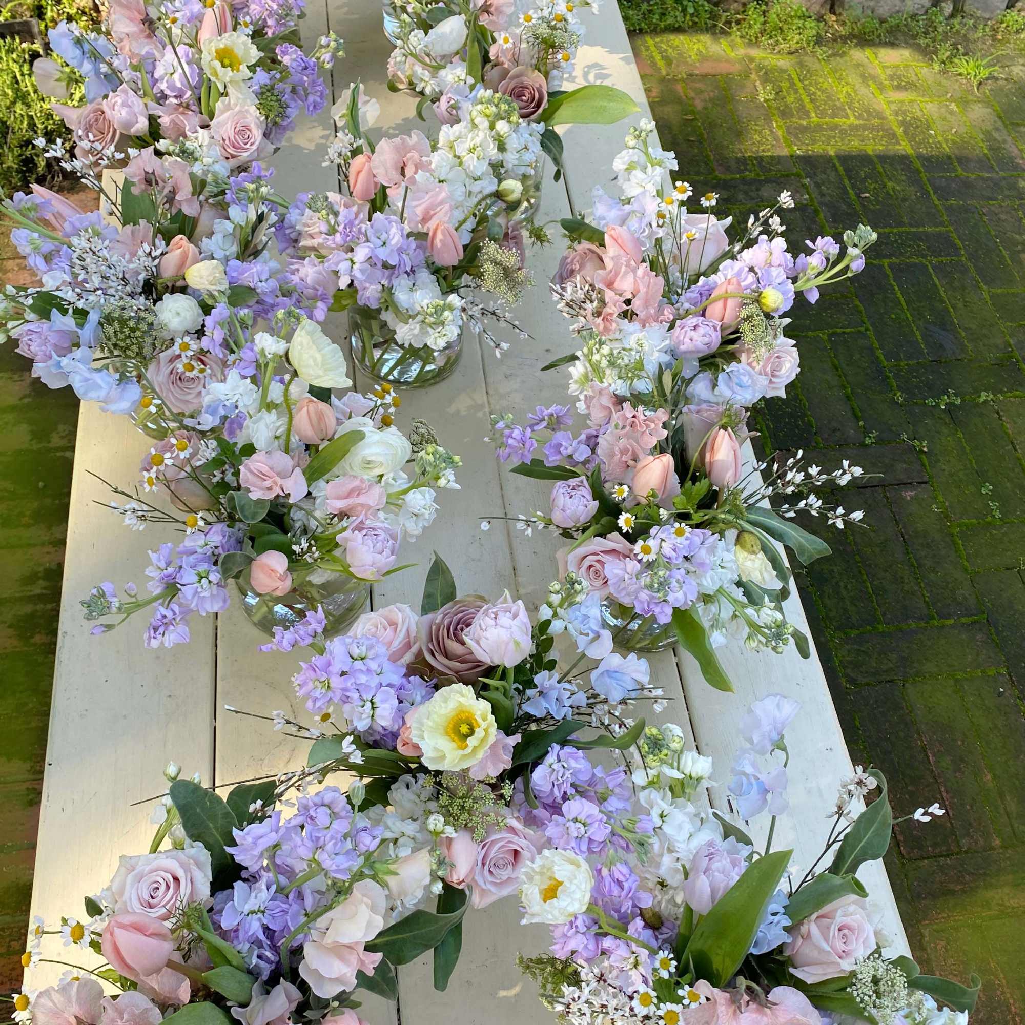 Pastel floral bouquets arranged in small glass vases on a long table outdoors.