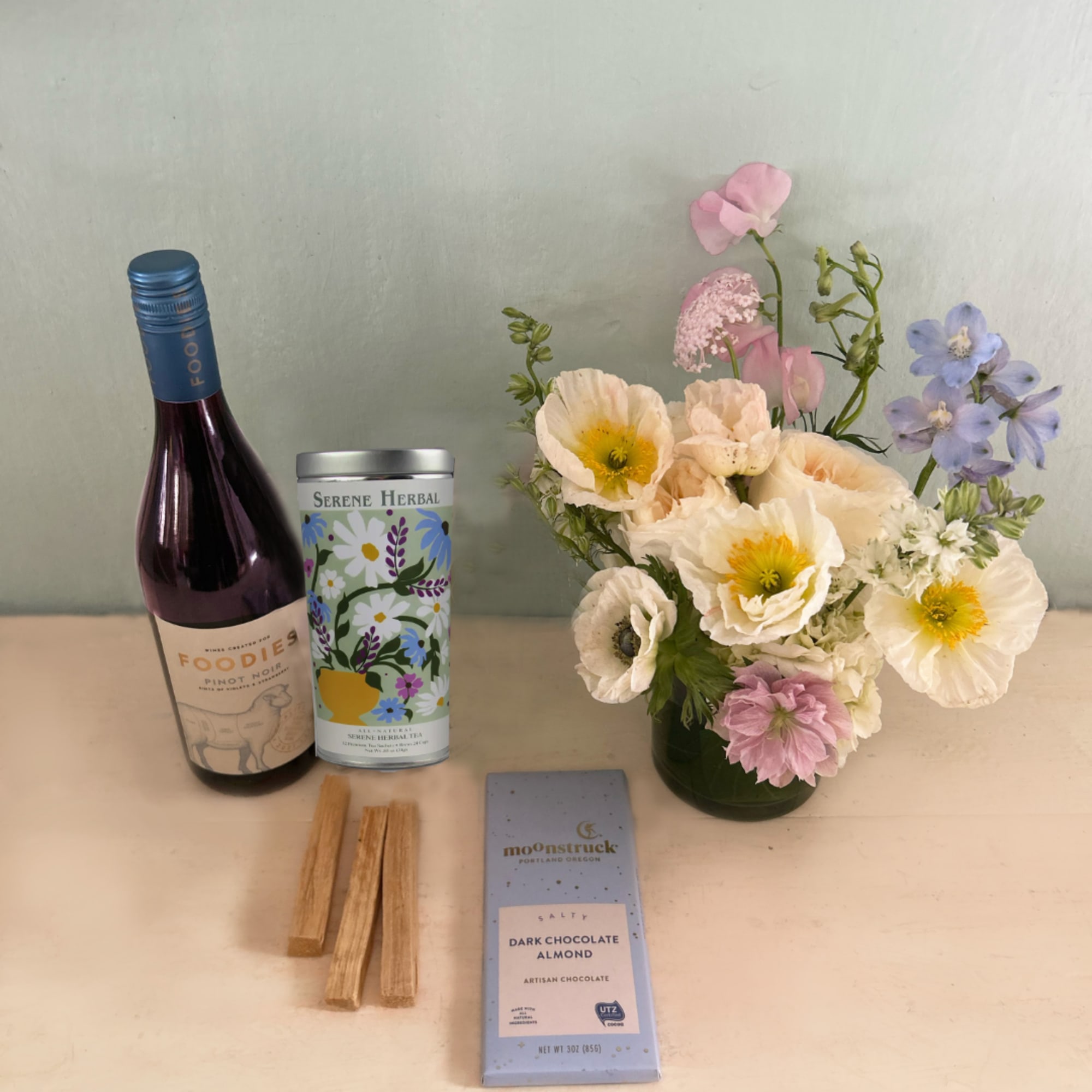 Flower arrangement in a green vase beside a wine bottle, tea tin, and chocolate bar