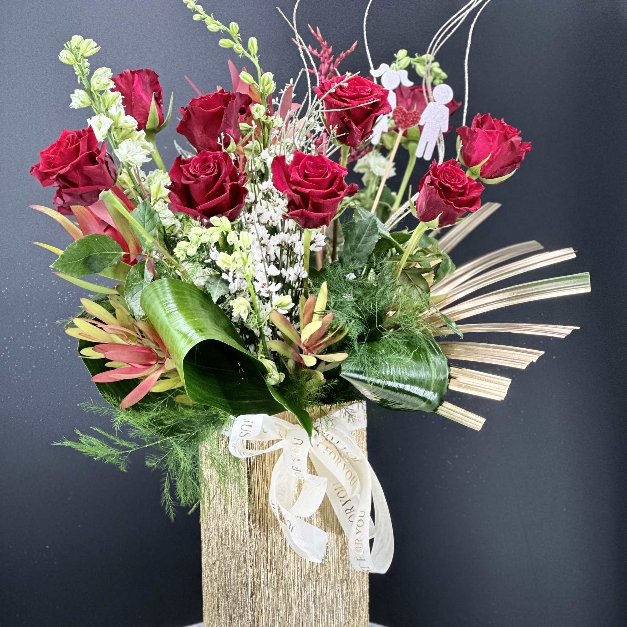 Red roses arranged in a gold vase with mixed greenery and decorative accents